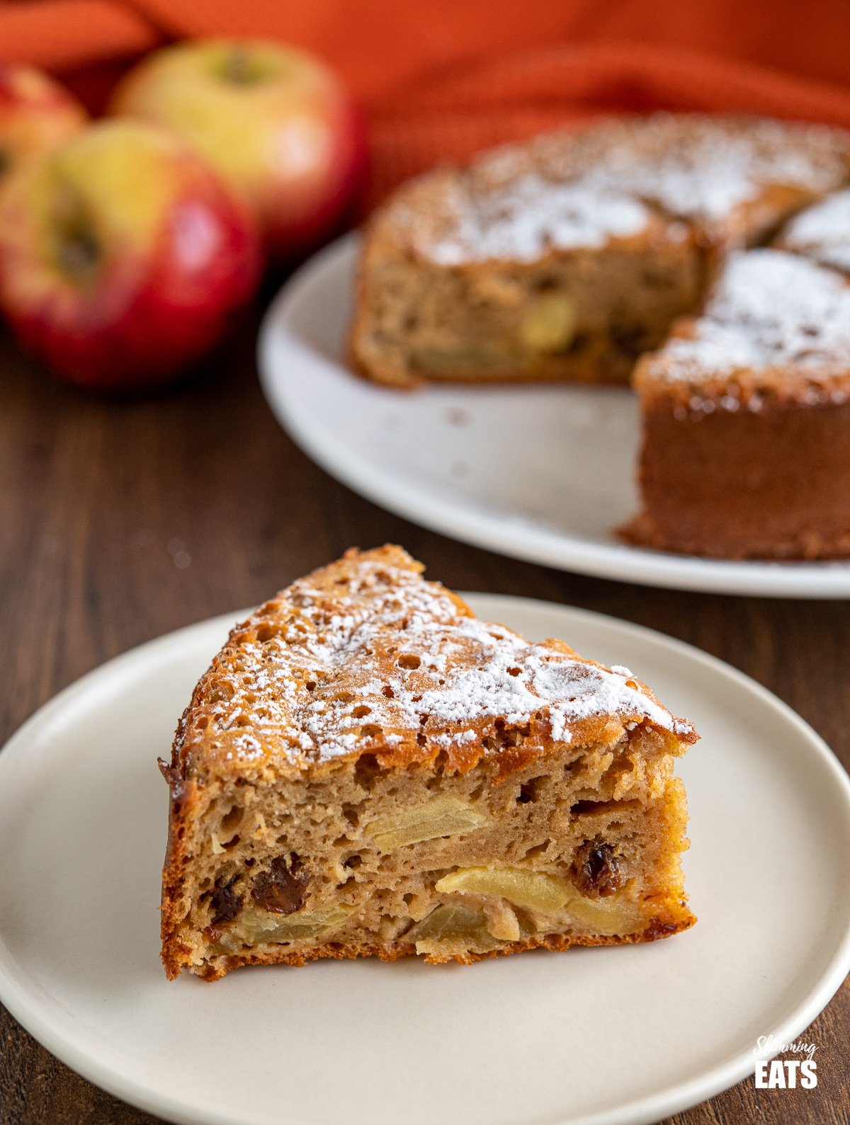 slice of apple sultana cake on white plate with cake and apples in background