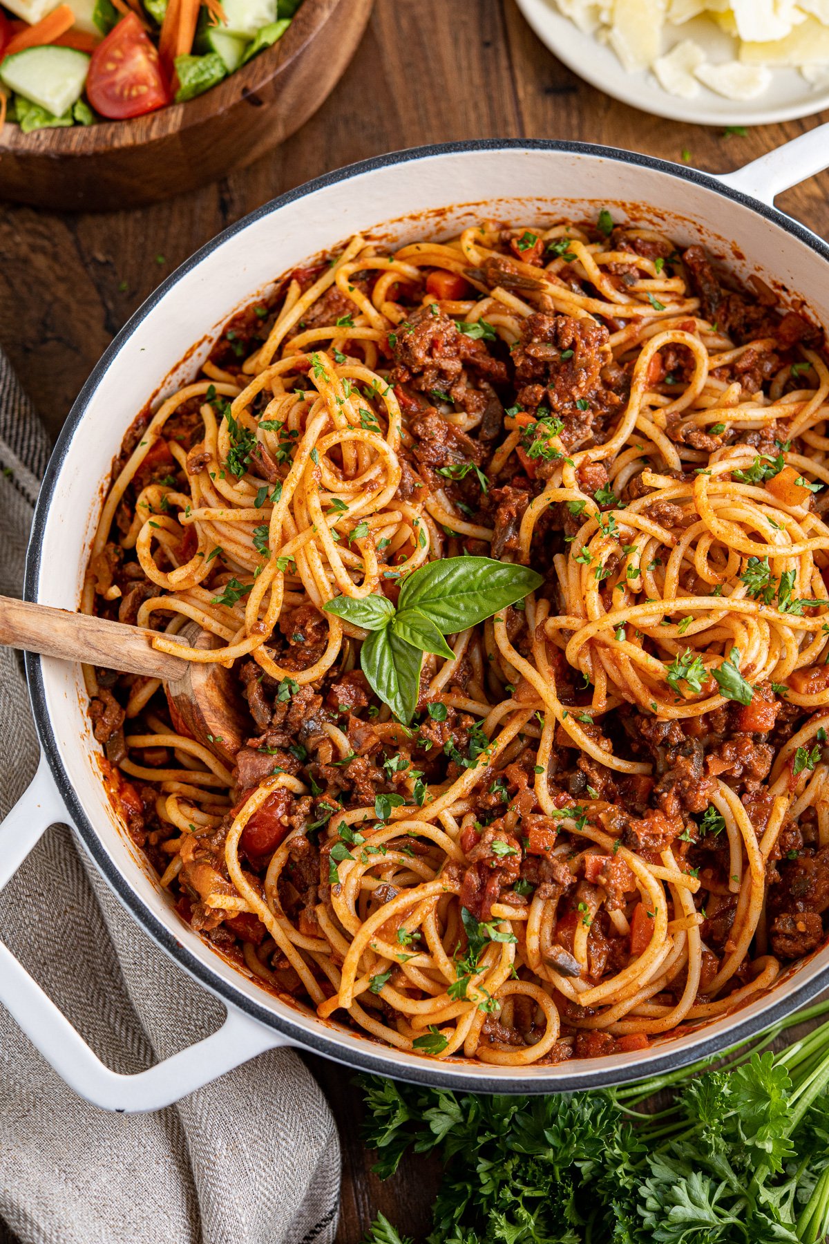 spaghetti bolognese mixed up in a white cast iron pan top with parsley and basil, bowl of salad and plate of parmesan in the background