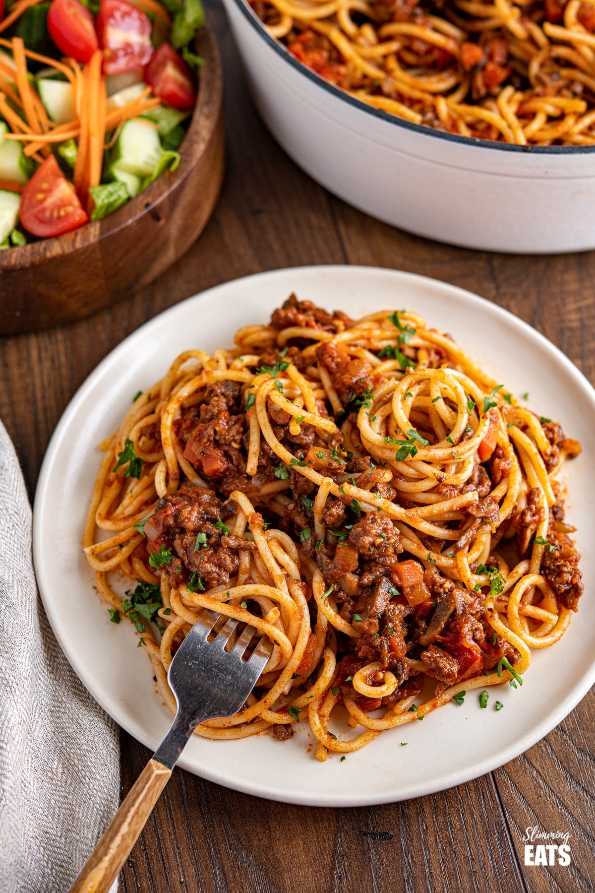 plated up spaghetti bolognese on small white plate with fork, pan and bowl of salad in background