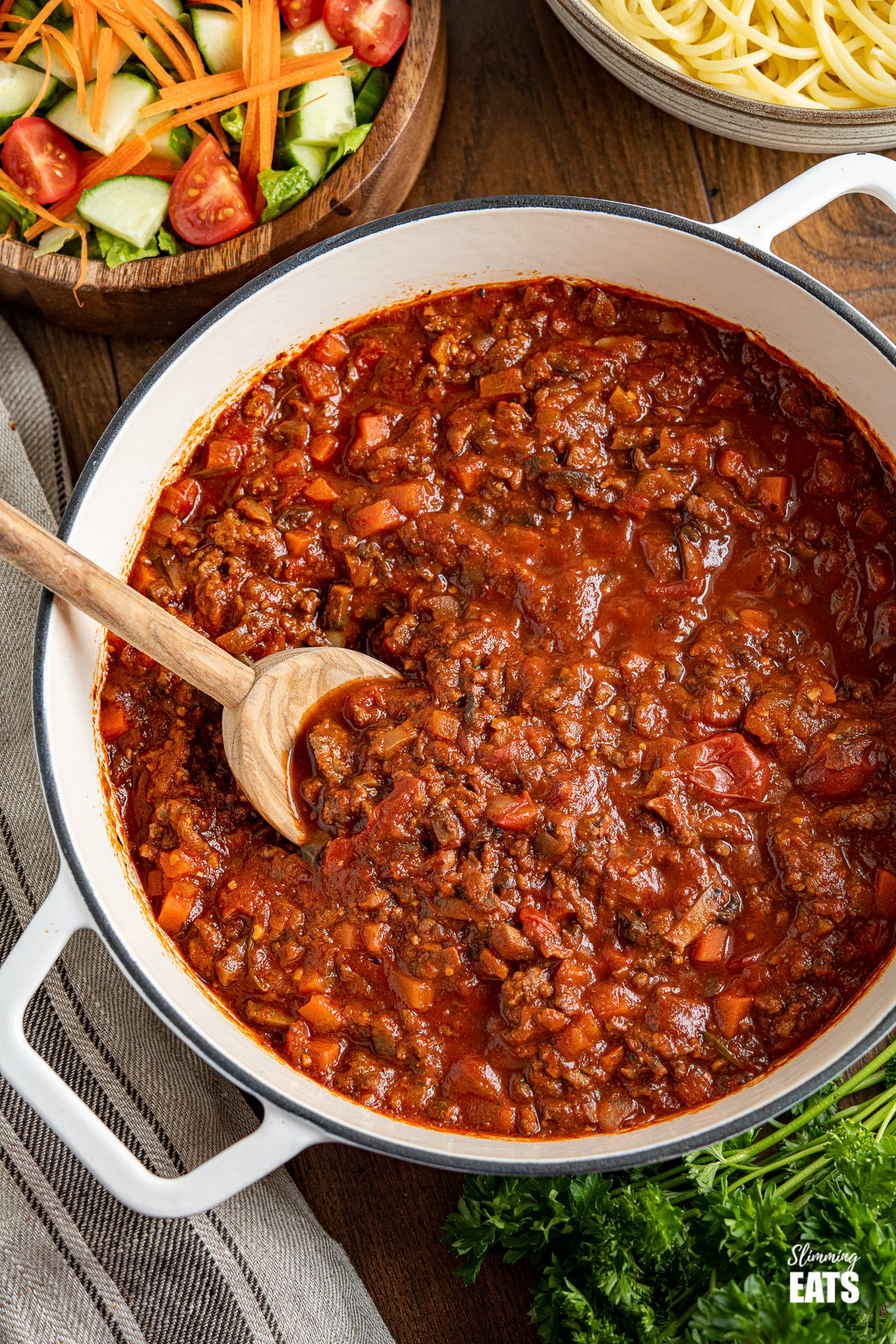 Bolognese meat sauce in white cast iron casserole pan with wooden bowl of salad and bowl of spaghetti in background