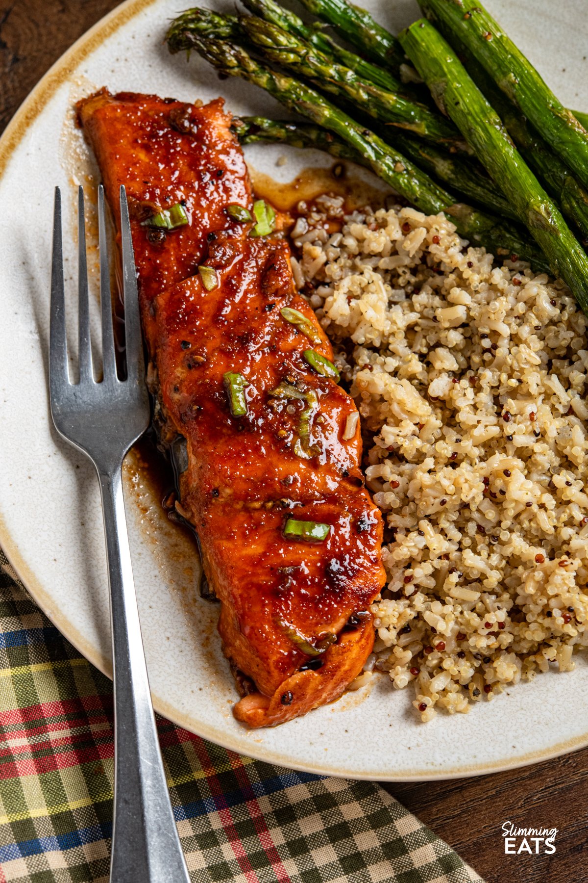 Delicious Maple Glazed Salmon – simple ingredients, amazing taste. Wild-caught salmon fillets are seasoned with sweet paprika, garlic powder, salt, and pepper, then baked in the oven with a rich, glossy glaze of maple syrup and low-sodium soy sauce. As the salmon bakes, the glaze caramelises beautifully, creating a deliciously savoury-sweet finish that enhances the natural flavour of the fish. Maple Glazed Salmon on a white plate with beige rim served with roasted asparagus and quinoa brown rice blend.