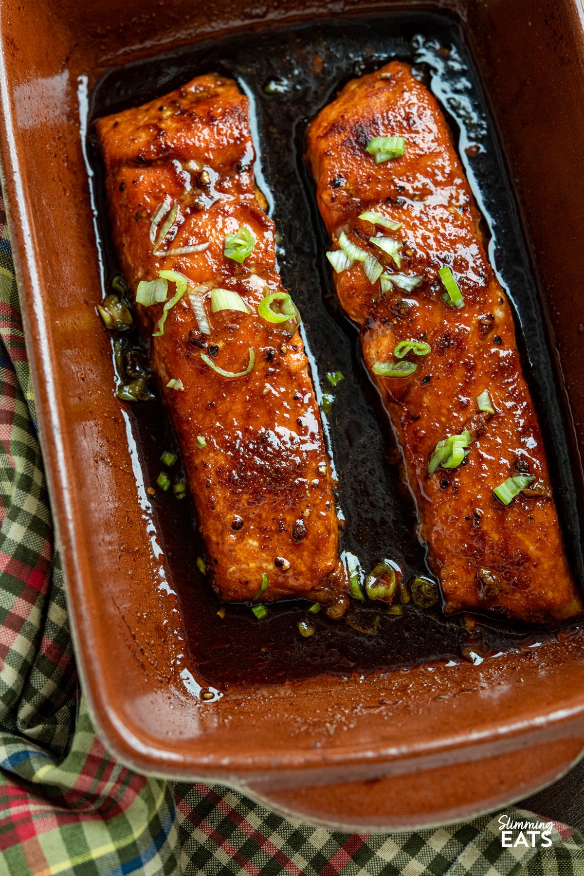 Delicious Maple Glazed Salmon – simple ingredients, amazing taste. Wild-caught salmon fillets are seasoned with sweet paprika, garlic powder, salt, and pepper, then baked in the oven with a rich, glossy glaze of maple syrup and low-sodium soy sauce. As the salmon bakes, the glaze caramelises beautifully, creating a deliciously savoury-sweet finish that enhances the natural flavour of the fish. close up of Maple Glazed Salmon in terracotta/beige baking surrounded