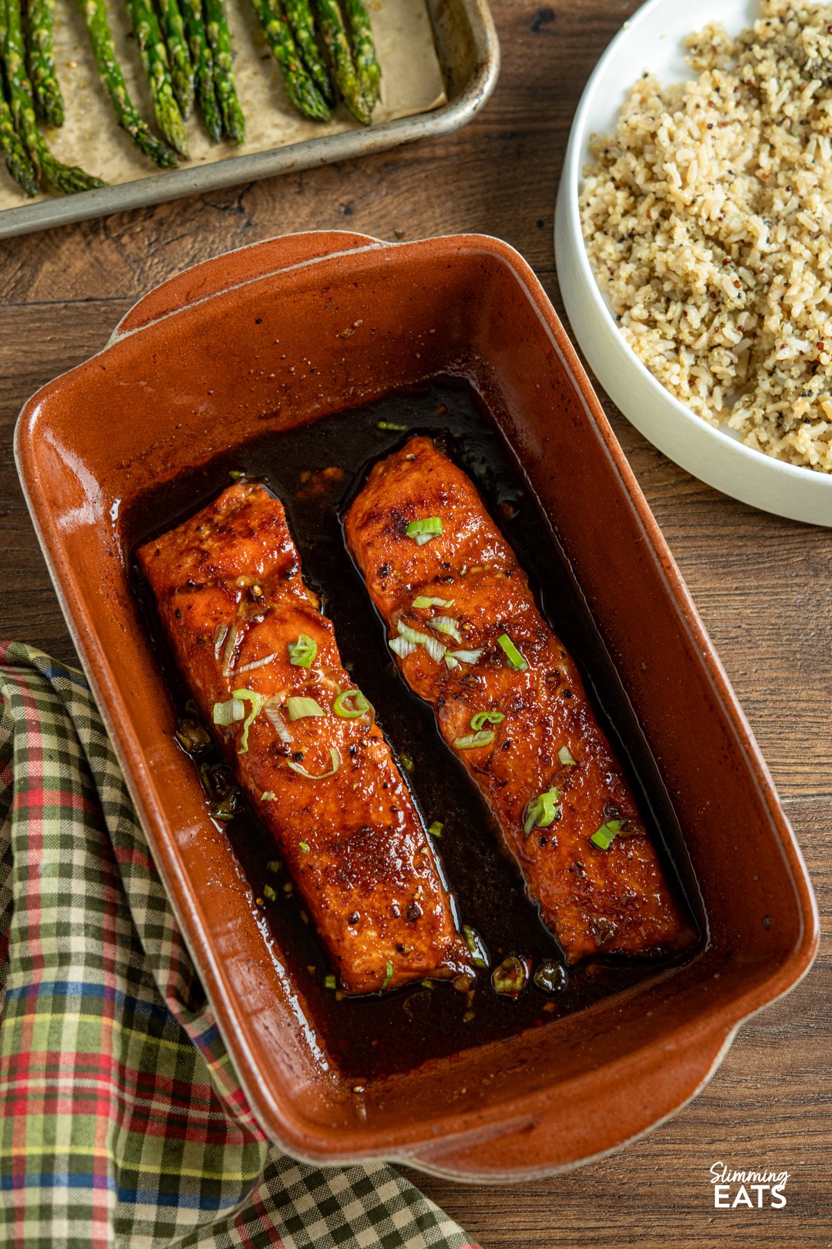 Delicious Maple Glazed Salmon – simple ingredients, amazing taste. Wild-caught salmon fillets are seasoned with sweet paprika, garlic powder, salt, and pepper, then baked in the oven with a rich, glossy glaze of maple syrup and low-sodium soy sauce. As the salmon bakes, the glaze caramelises beautifully, creating a deliciously savoury-sweet finish that enhances the natural flavour of the fish. Maple Glazed Salmon in terracota/beige baking surrounded by sides of roasted asparagus and quinoa brown rice mix.