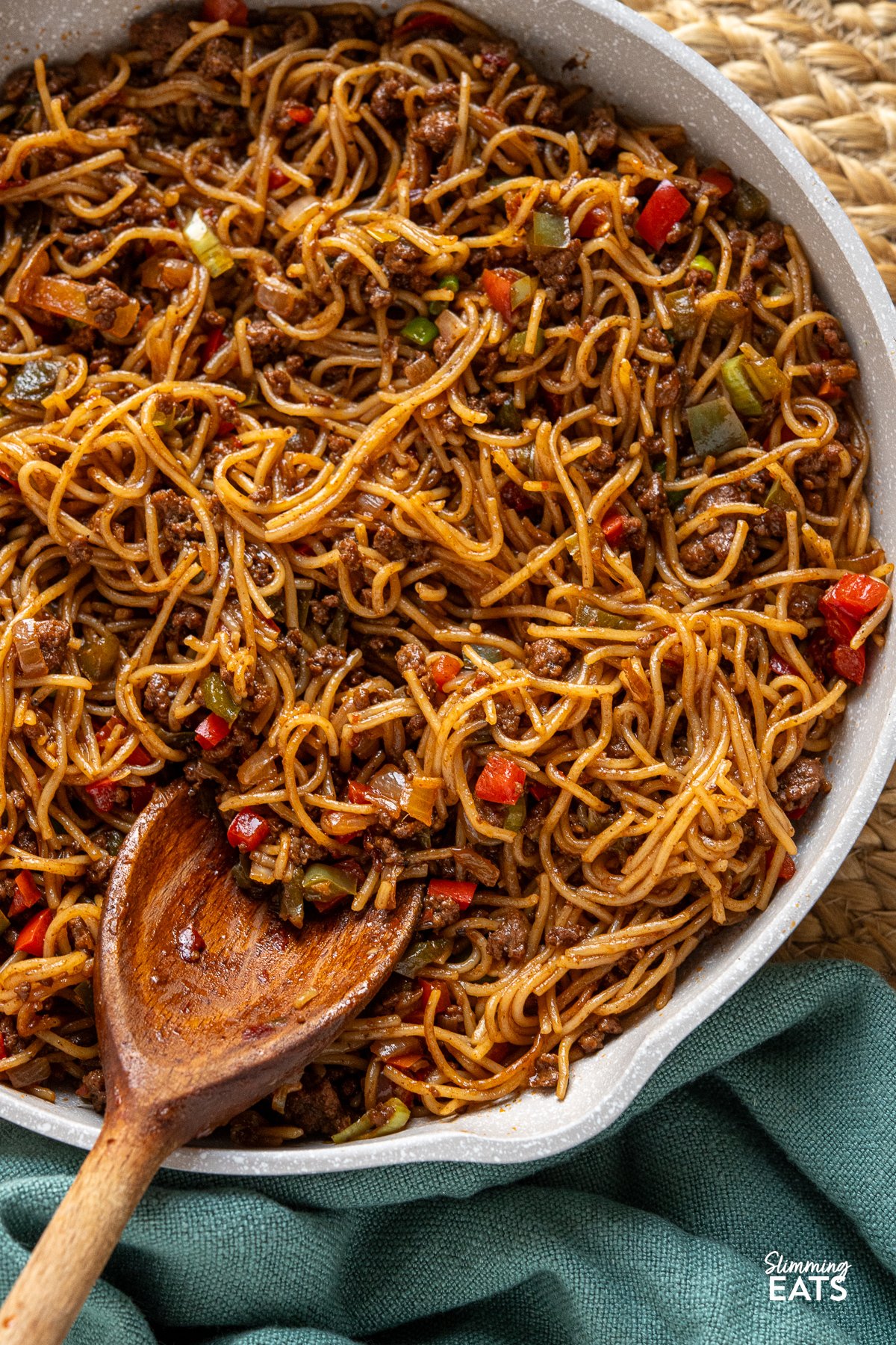 Chilli beef noodles cooking in a beige non-stick frying pan with a wooden handle. A wooden spoon is placed in the pan.