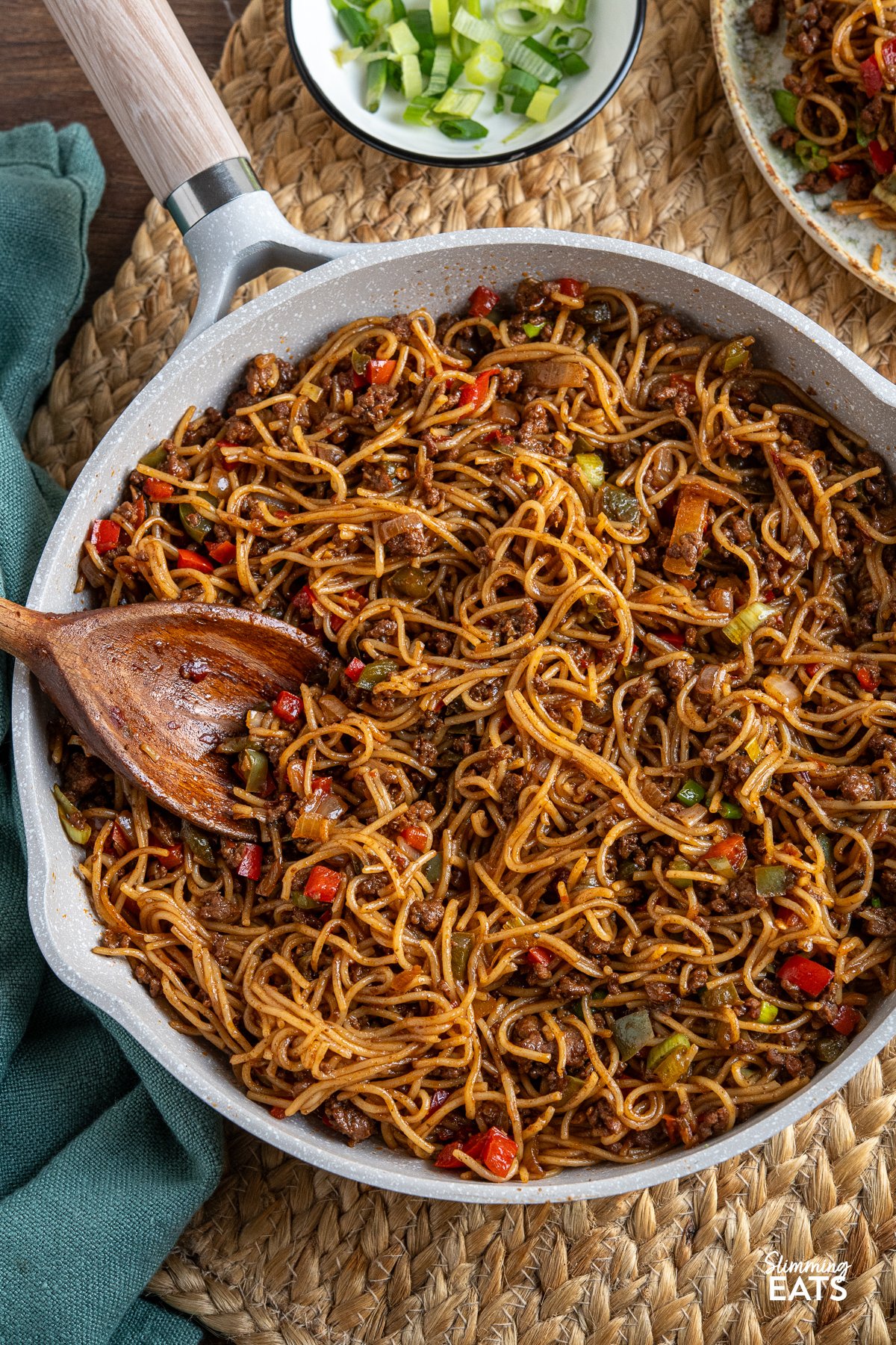 Chilli beef noodles cooking in a beige non-stick frying pan with a wooden handle. A wooden spoon is placed in the pan.