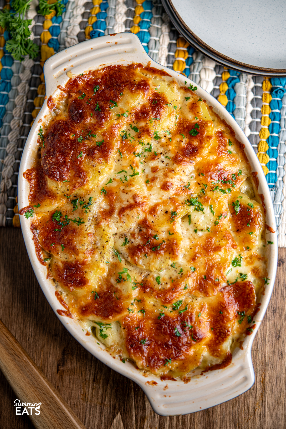 cheesy leek and potato bake in a le Creuset oval dish on wooden board with plates and blue, yellow, creamy and grey coloured fabric placemat