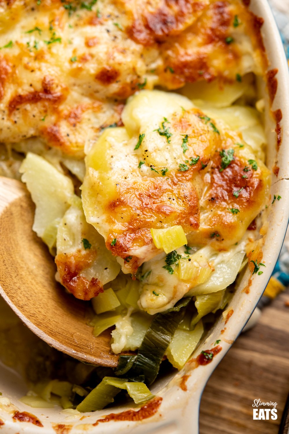 Delicious Cheesy Leek and Potato Bake - easy to make and perfect to enjoy as a side dish or even a main course. Gluten Free, Vegetarian, Slimming World and Weight Watchers friendly wooden spoon dishing up leek and potato bake from oval baking dish