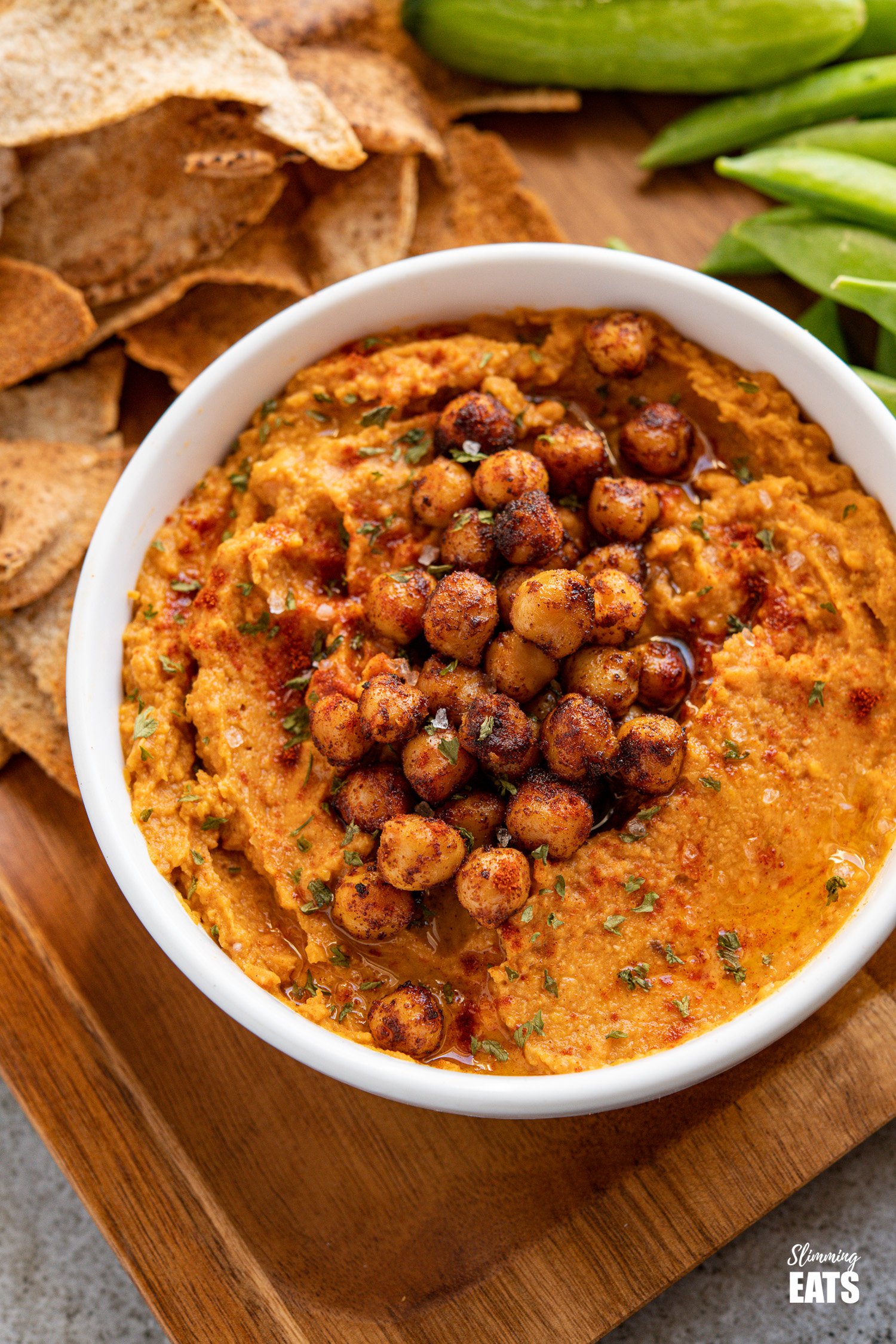 close up of roasted sweet potato hummus topped with toasted chickpeas in a white bowl on wooden tray with pita chips and vegetable crudites