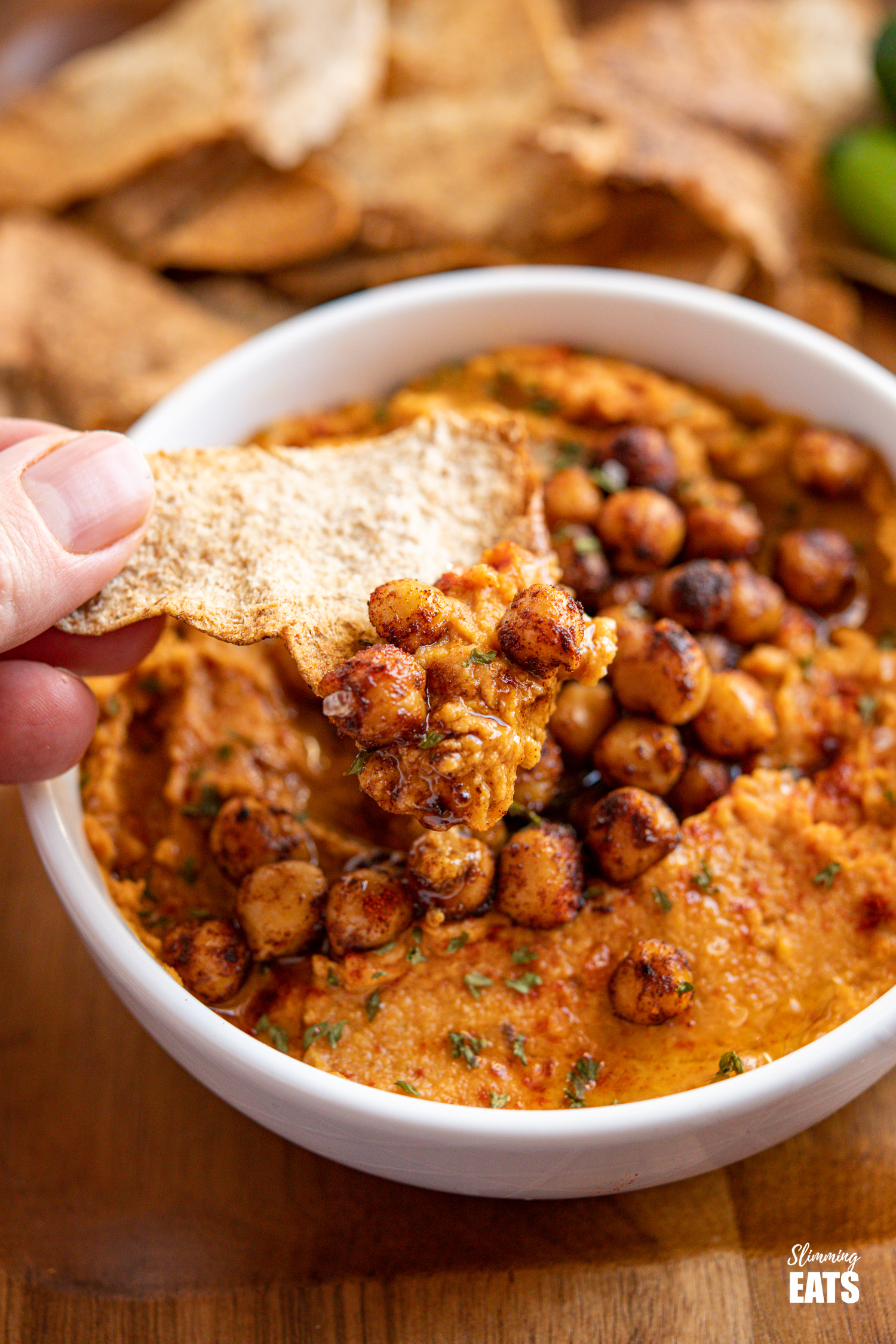 pita chip scooping roasted sweet potato hummus topped with toasted chickpeas from a white bowl