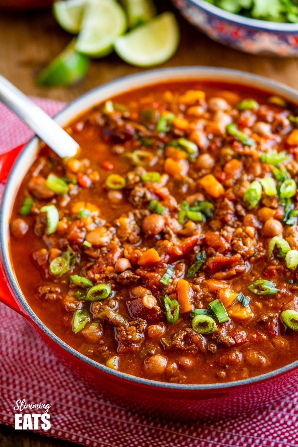 close up of beef and six bean chilli in red le creuset dutch oven with lime slices and salad in background