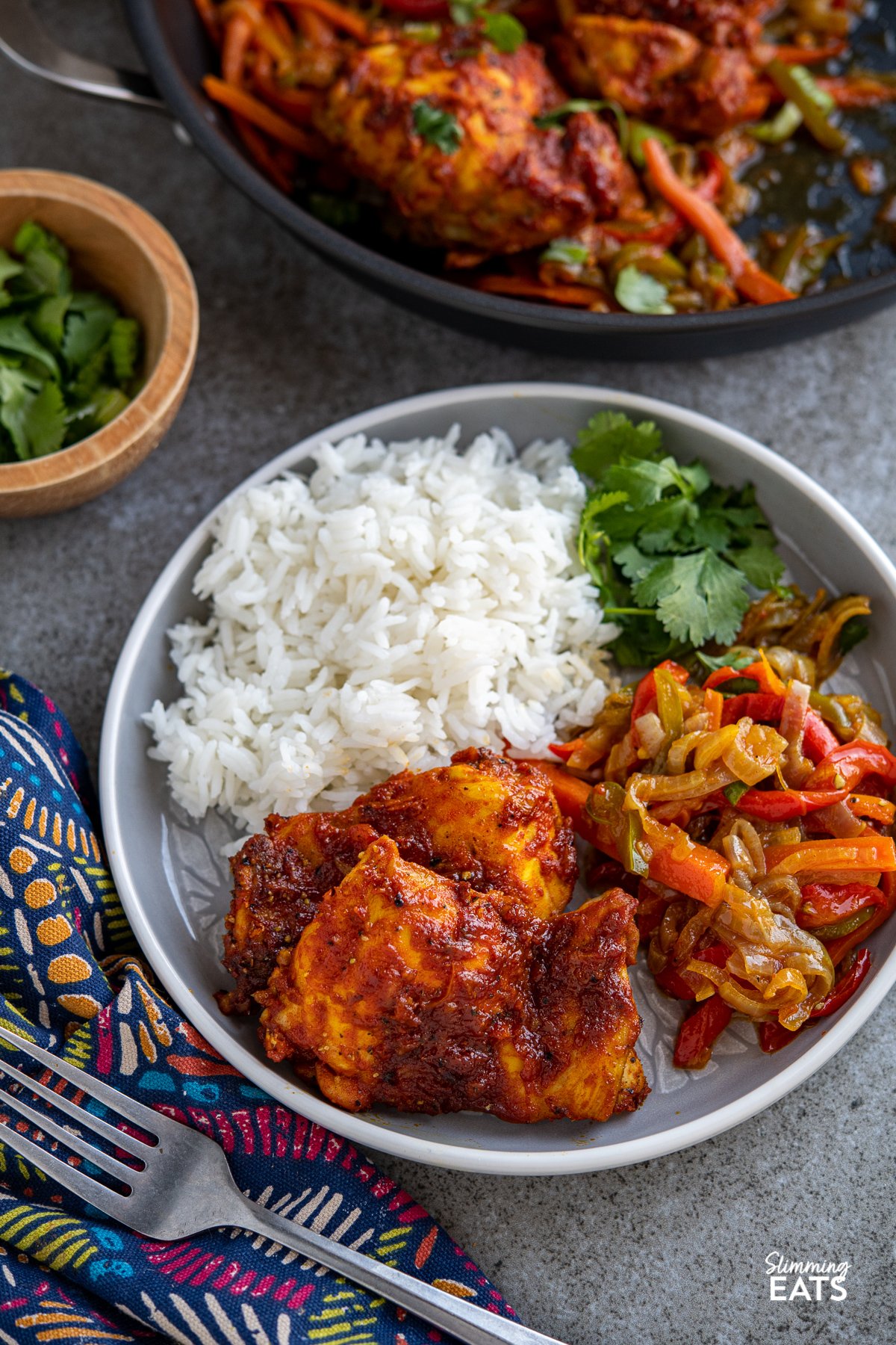 oven baked fusion chicken thighs on grey white rimmed plate with baked vegetables rice and cilantro, frying pan in background and bowl of cilantro