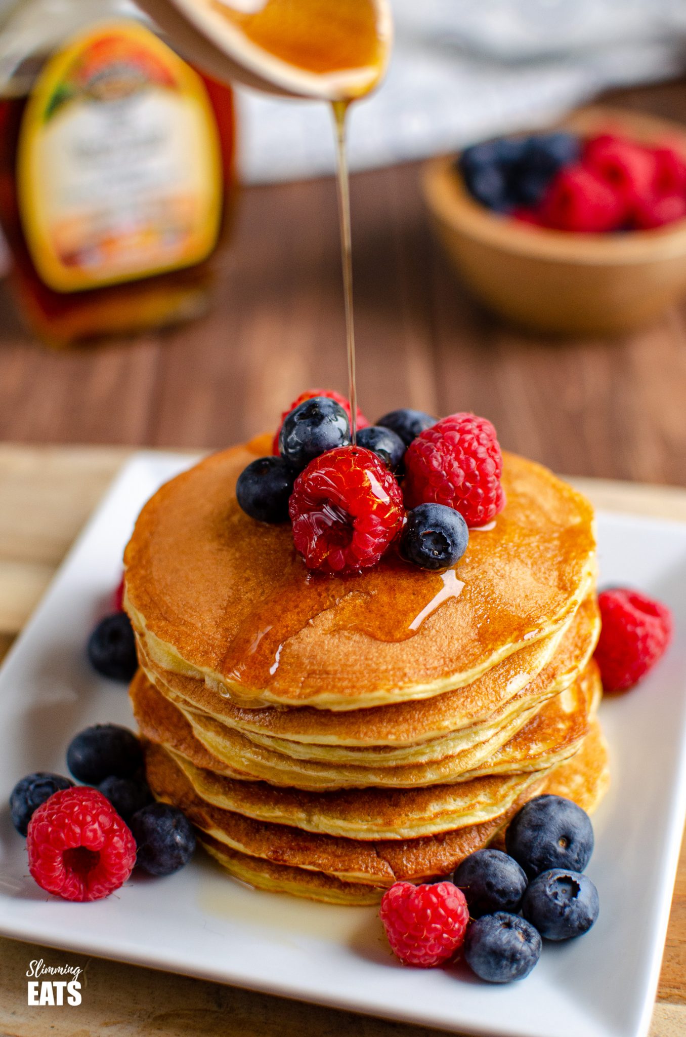 maple syrup being drizzled on American Style Pancakes on white plate with fruit, maple syrup bottle in background