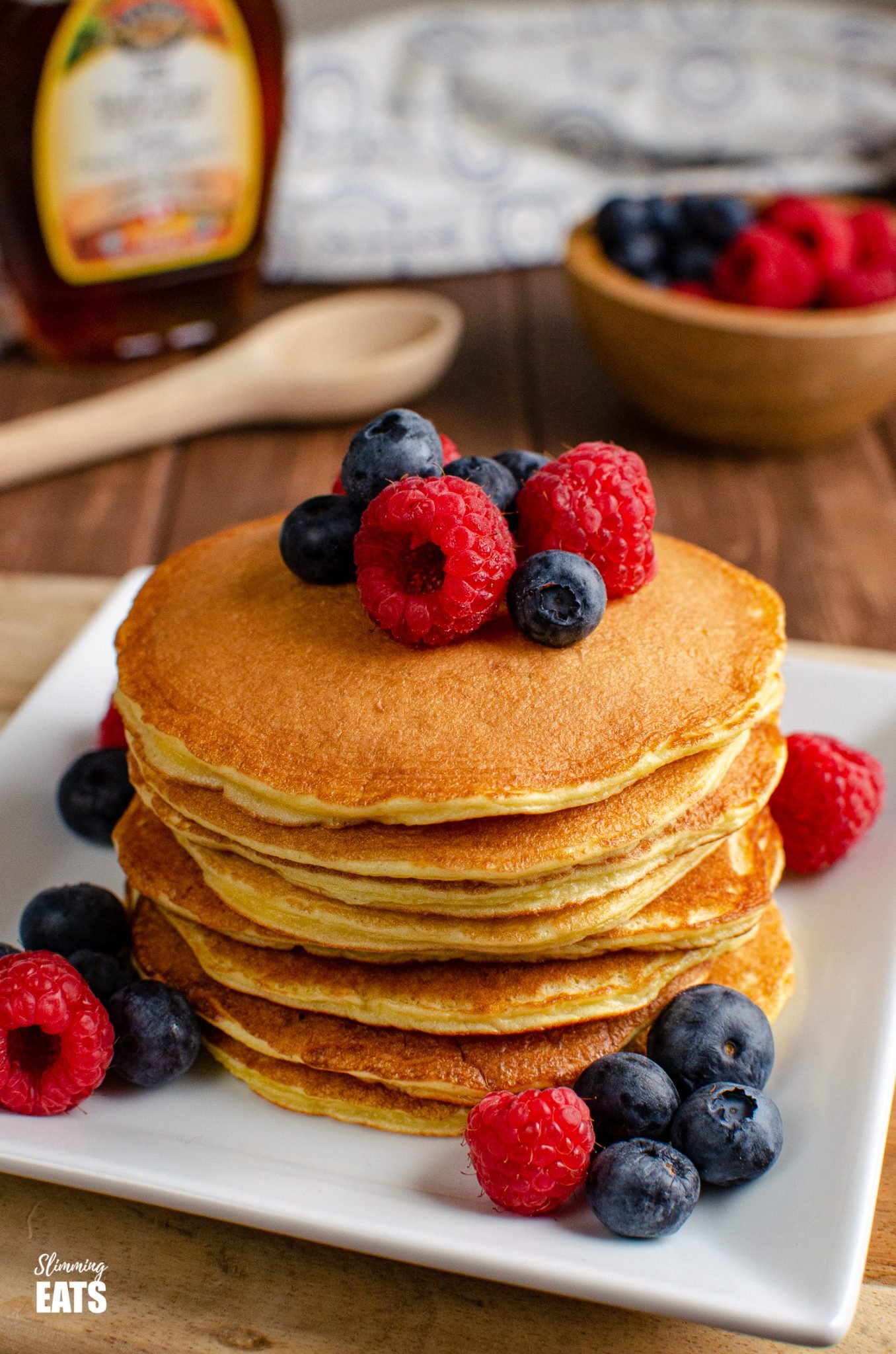 American Style Pancakes on white plate with fruit, maple syrup bottle in background