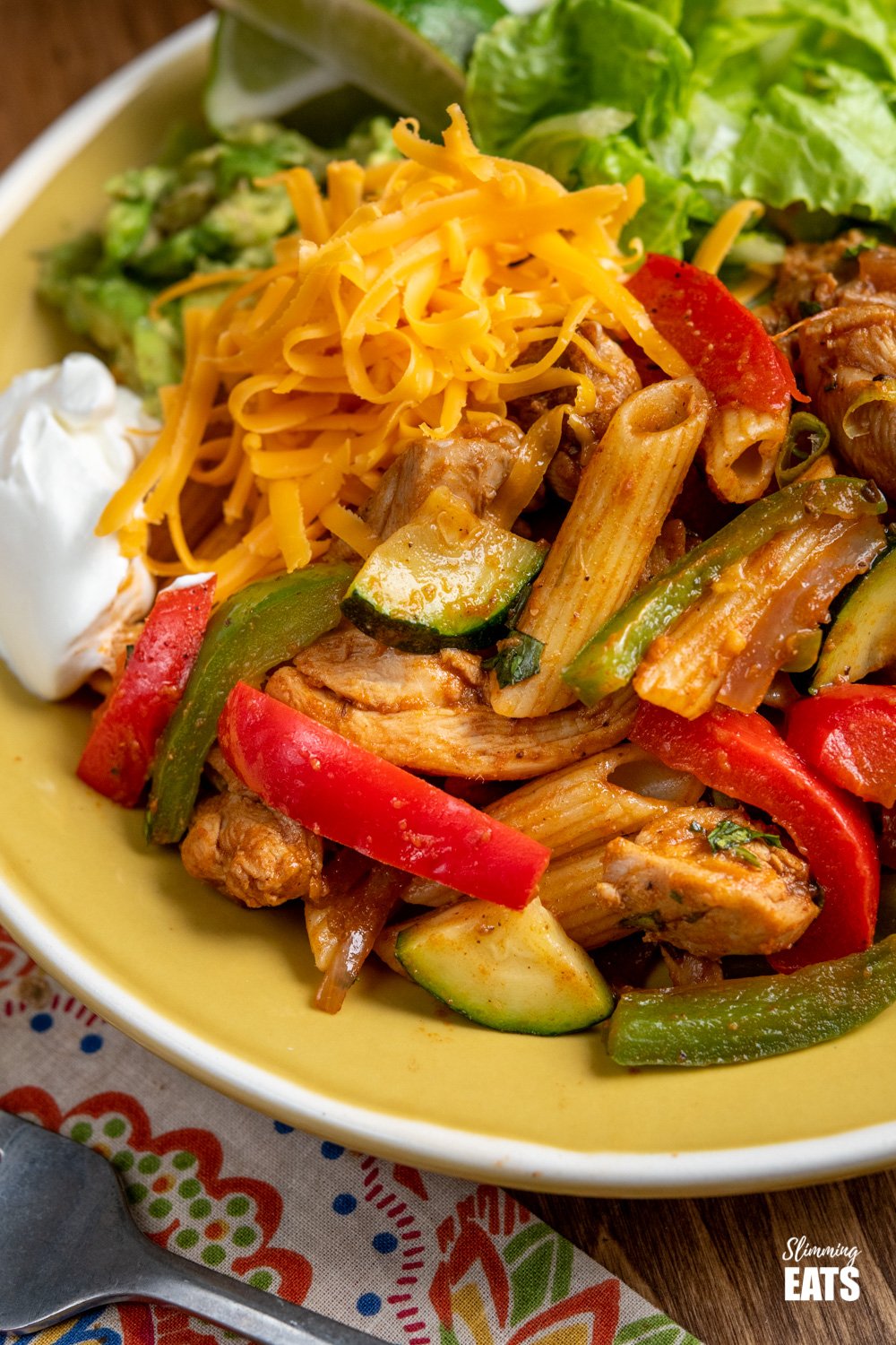 close up of serving of Fajita Chicken Pasta on a yellow plate with lettuce, cheddar, avocado, soured cream, and lime slices