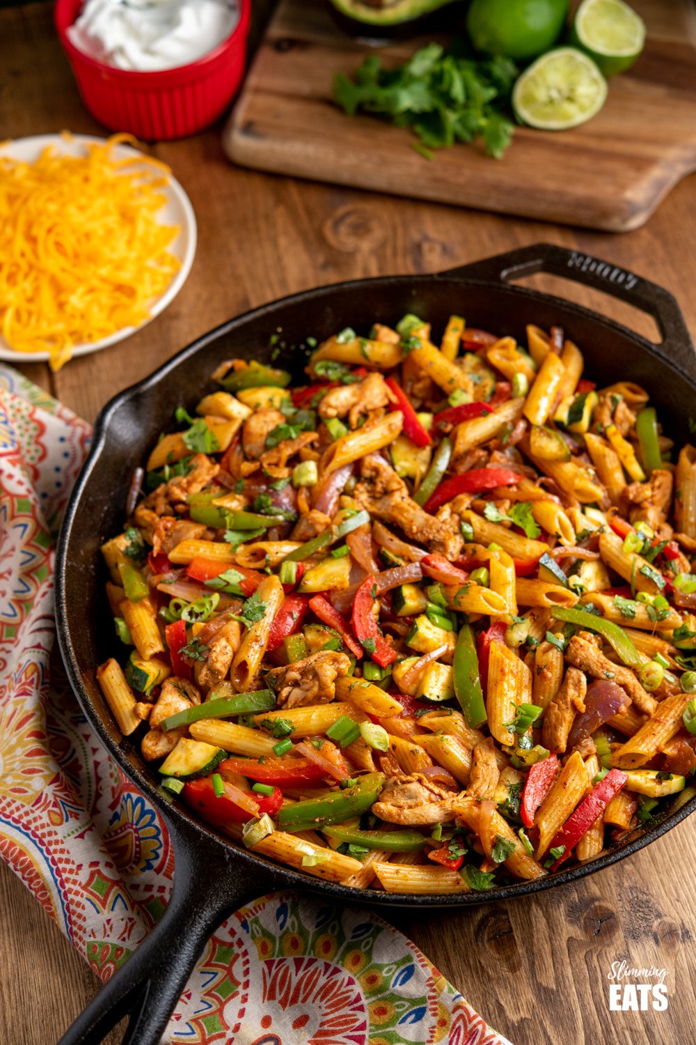 Fajita Chicken Pasta in a lodge cast iron skillet with cheddar, soured cream, limes and avocado in background