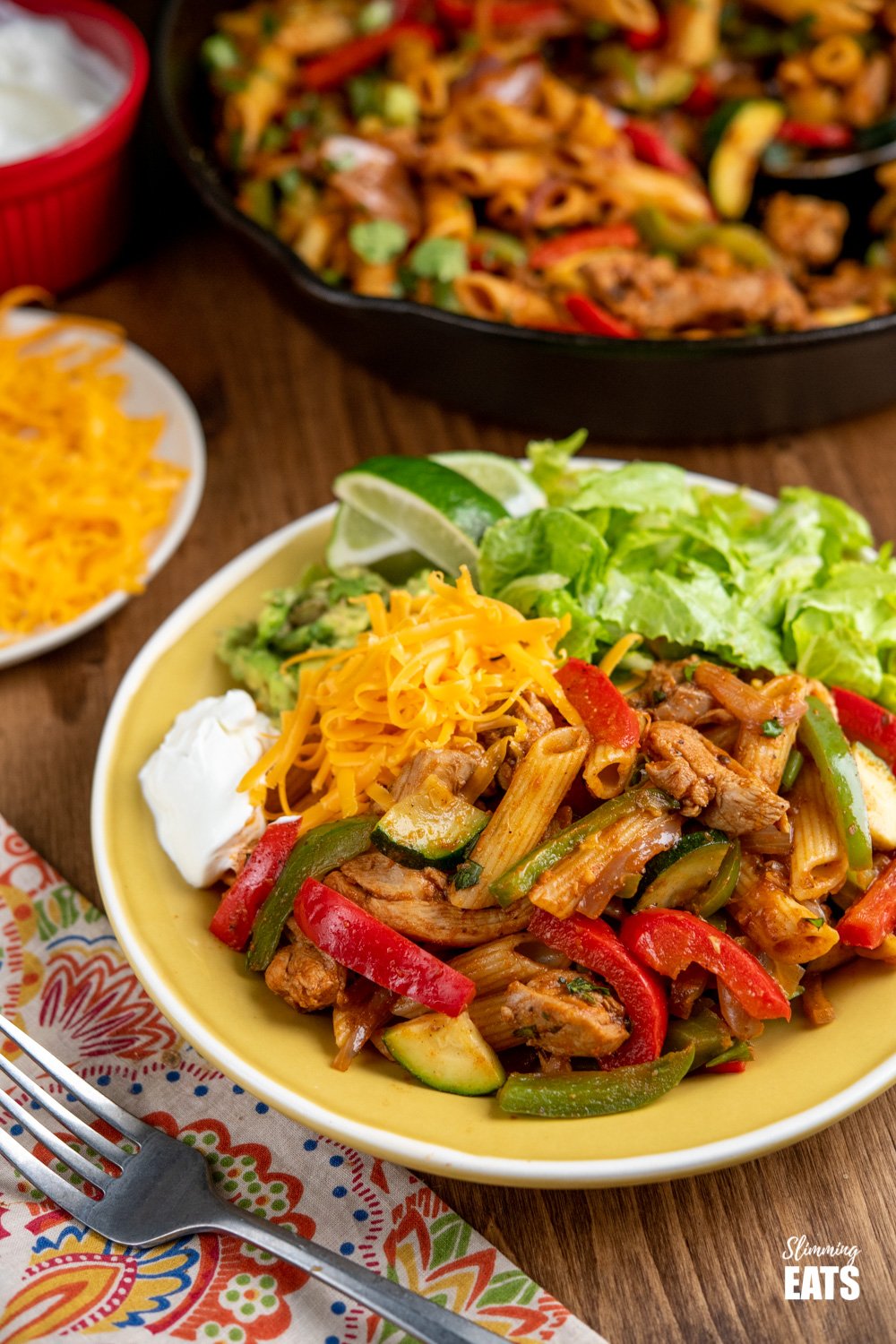 serving of Fajita Chicken Pasta on a yellow plate with lettuce, cheddar, avocado, soured cream, and lime slices