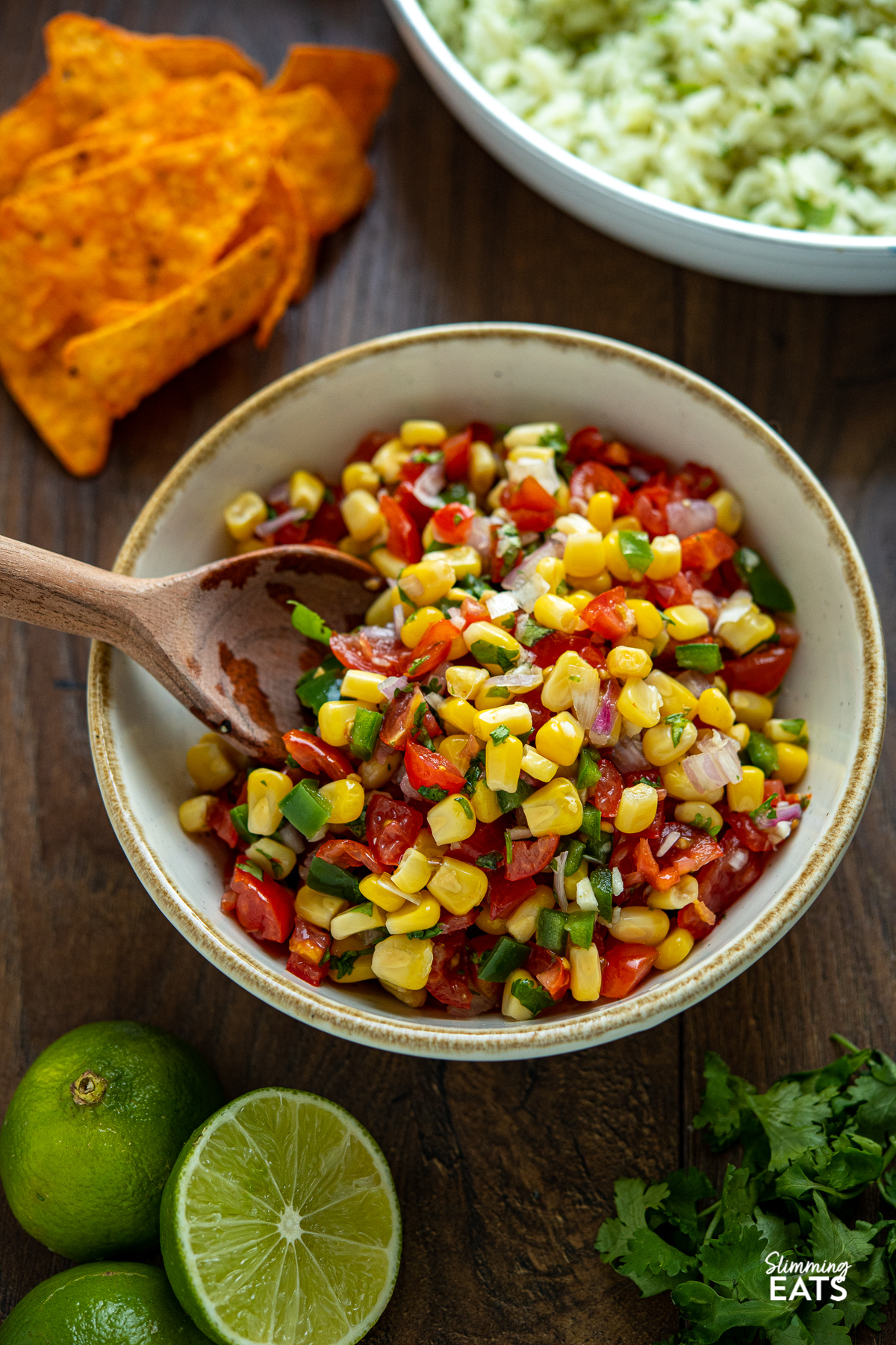 Easy Corn and Tomato Salsa in white bowl with light brown rim and wooden spoon with scattered limes and cilantro
