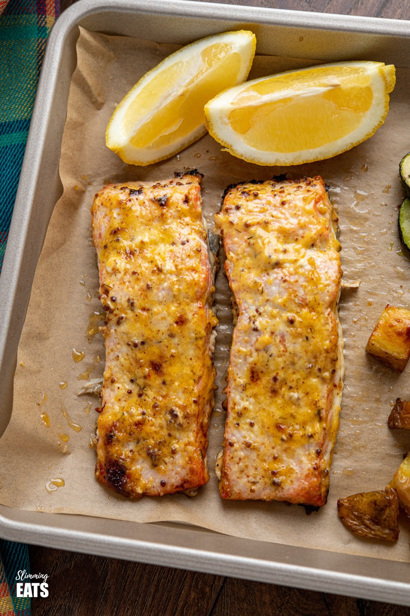 close up of Oven Baked Mustard Salmon Fillets on parchment lined tray with potatoes, zucchini and lemon wedge