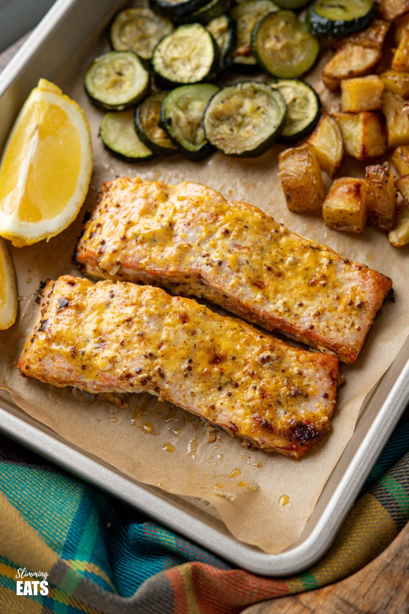 Oven Baked Mustard Salmon Fillets on parchment lined tray with potatoes, zucchini and lemon wedge