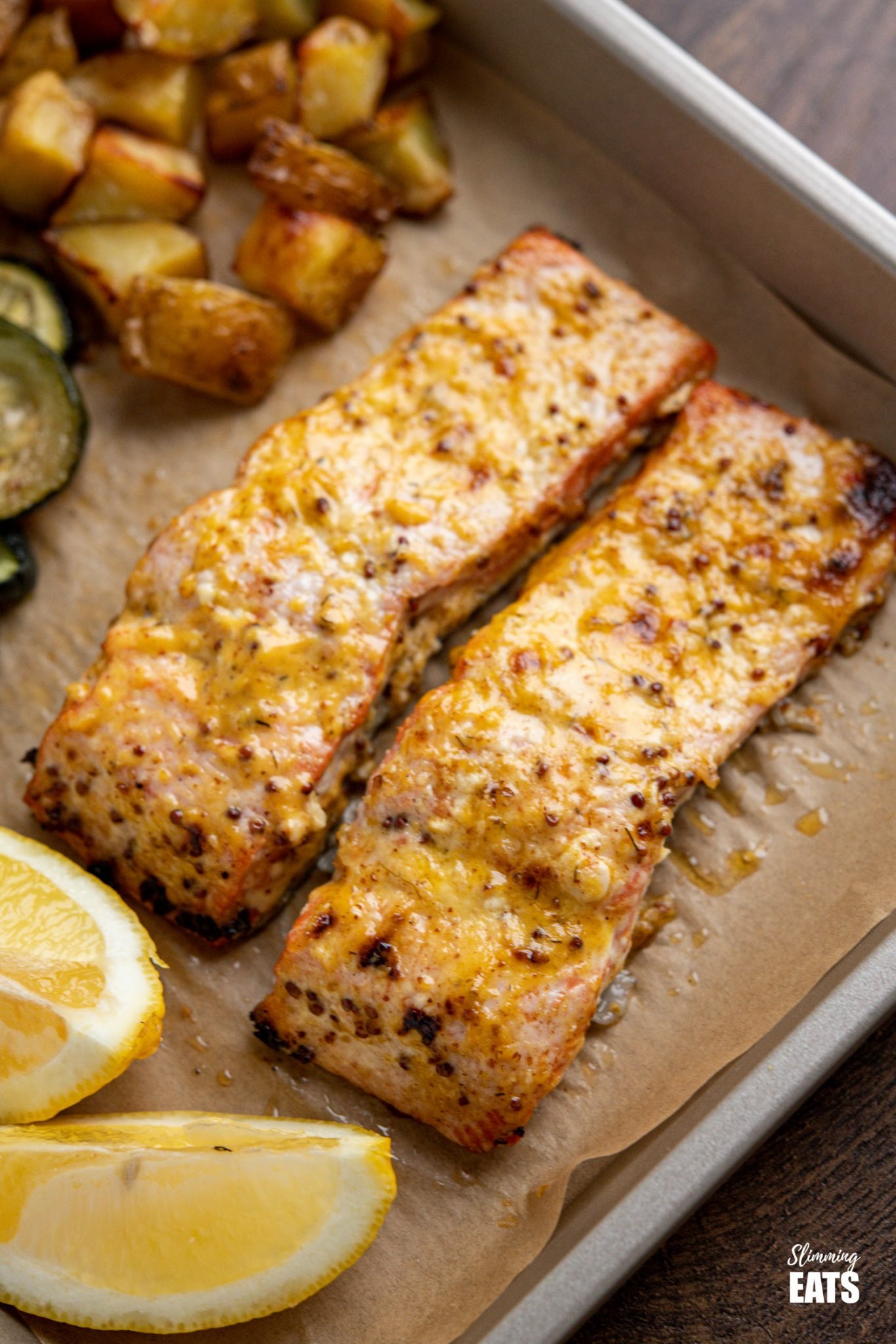 Oven Baked Mustard Salmon Fillets on parchment lined tray with potatoes, zucchini and lemon wedge