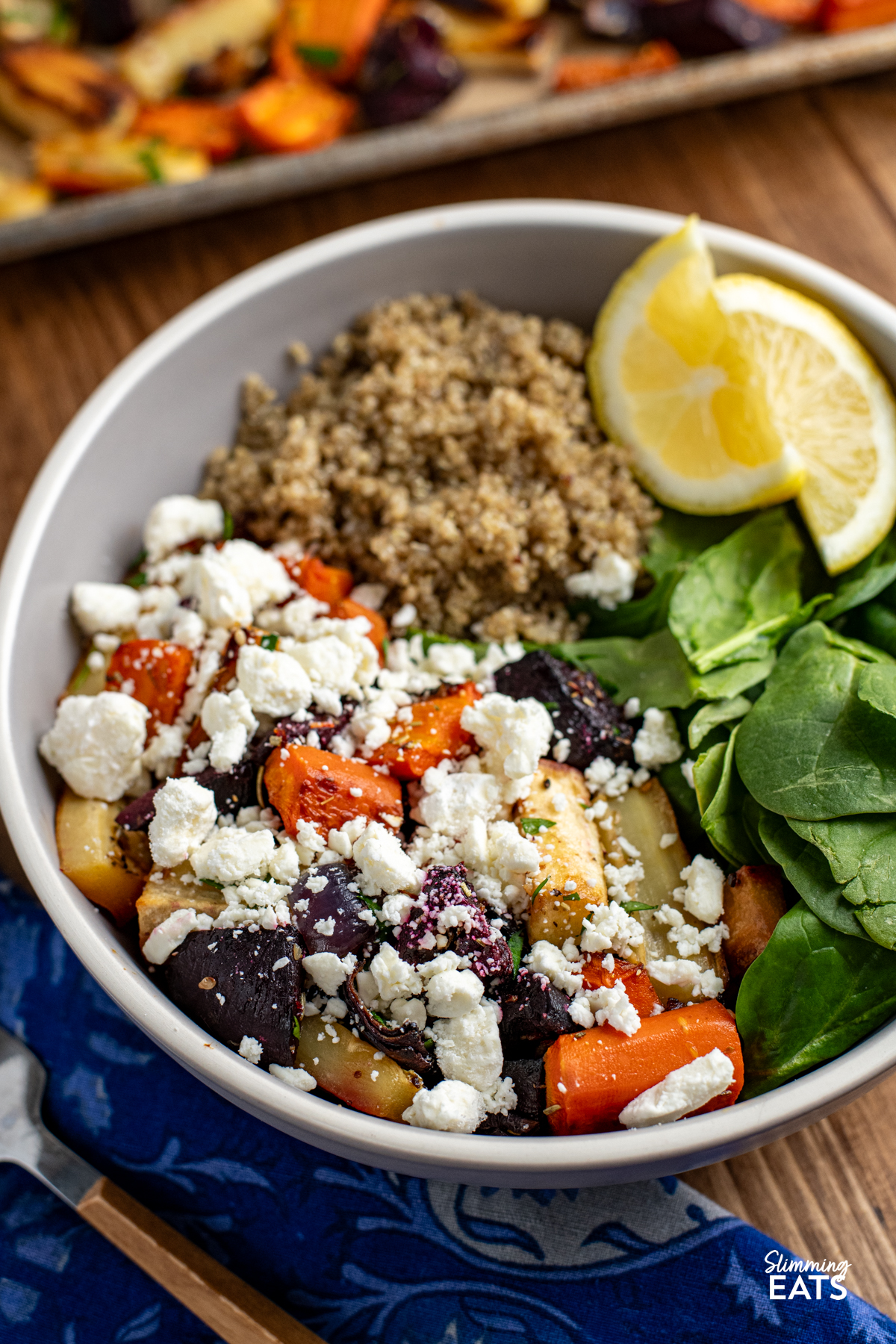 roasted parsnips, carrot, beets in a grey bowl topped with crumbled feta and served with cooked quinoa, baby spinach and lemon wedges, fork on side