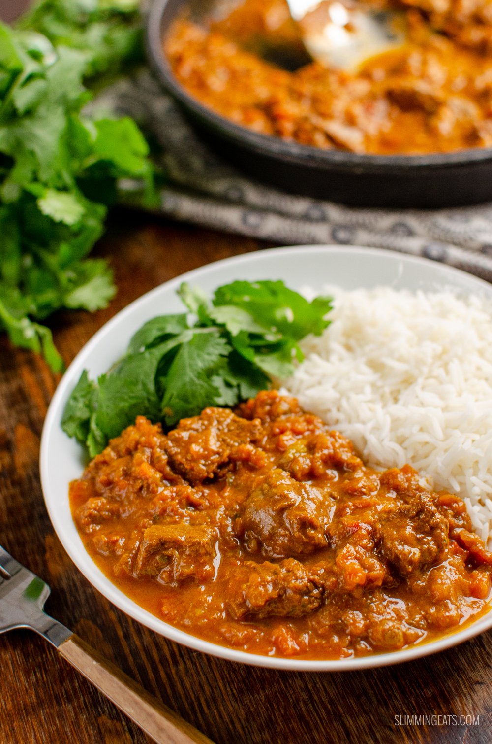 bombay beef curry in white bowl with rice and coriander, pan visible in background