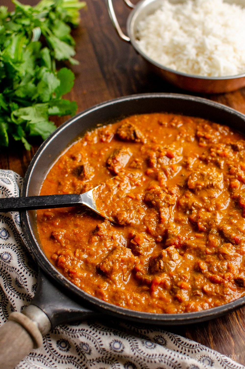 Bombay beef curry in a pan with rice and coriander in background