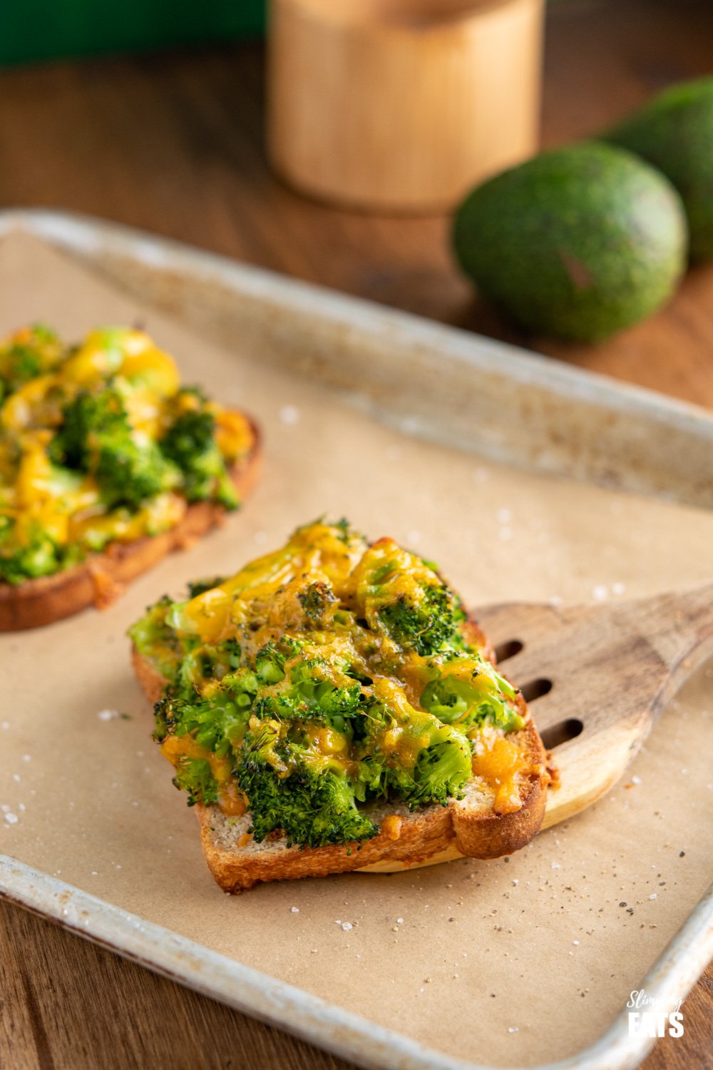 cheesy broccoli avocado toasts on a parchment lined baking tray with wooden spatula