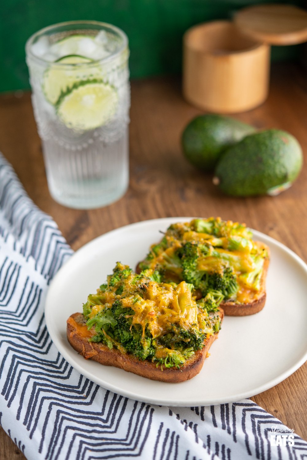 cheesy broccoli avocado toasts on white plate with a glass of lime water and avocados in background