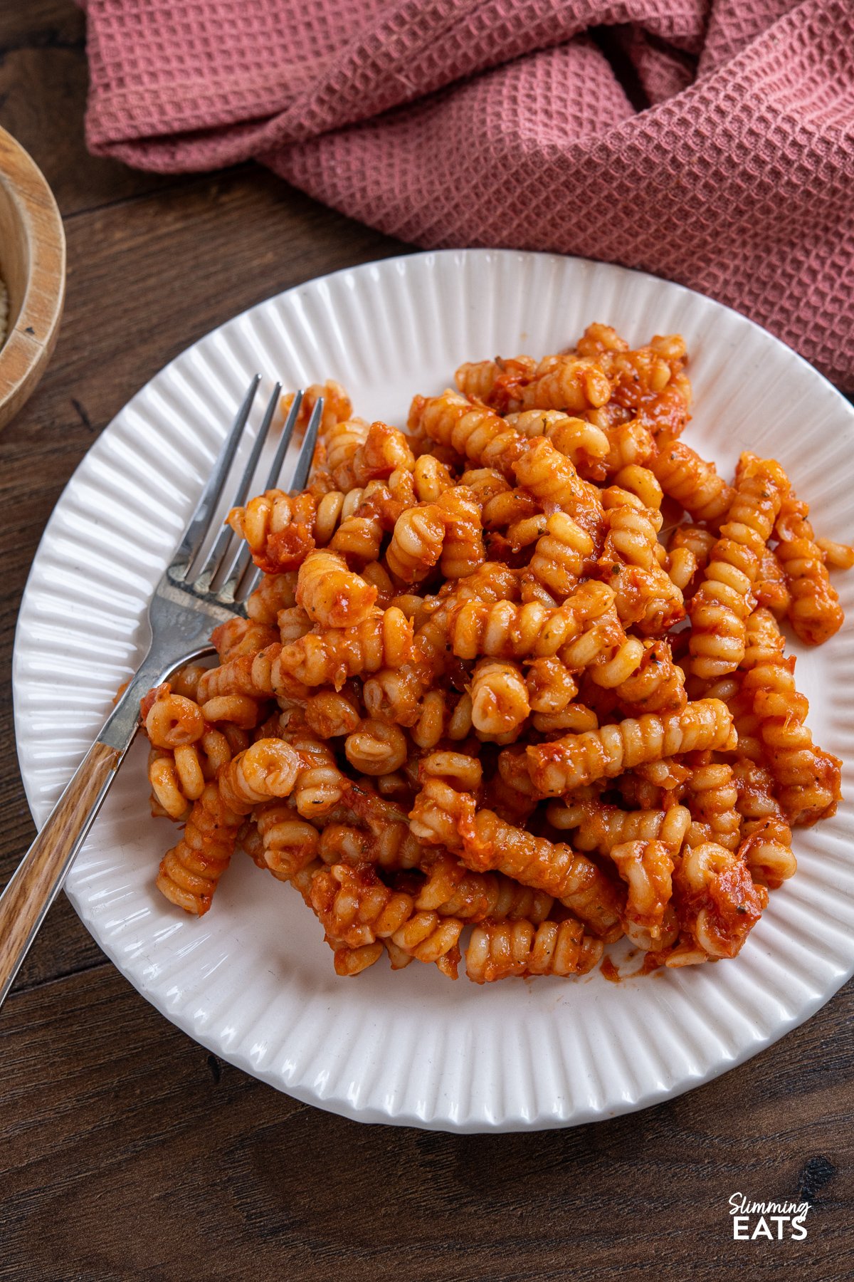 Pasta on a white/cream plate covered in the roasted tomato and garlic sauce, with a small wooden bowl of freshly grated Parmesan cheese in the background.