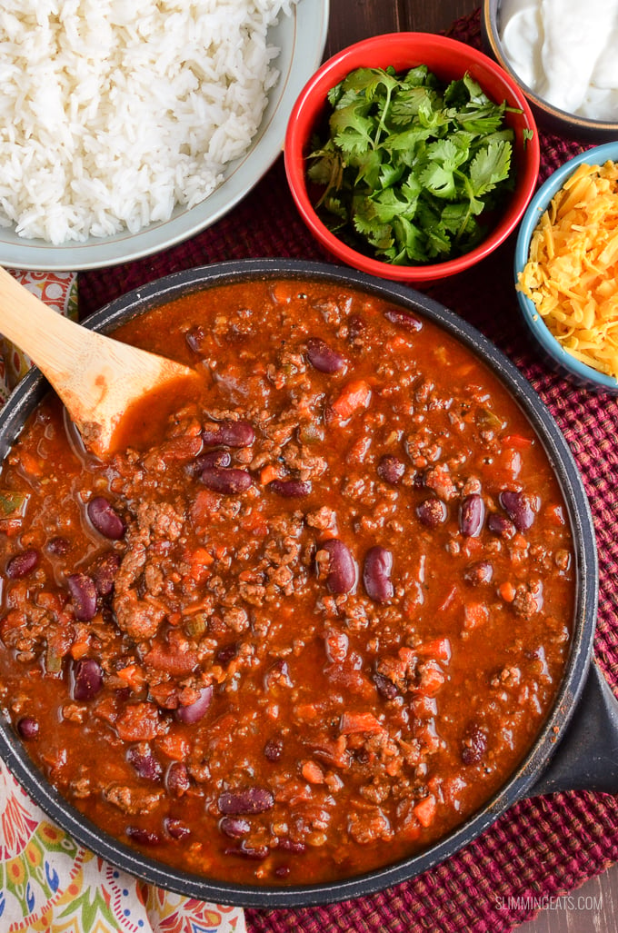 Chilli Con Carne in pan with wooden spoon and bowls with rice, cilantro, cheddar and soured cream.