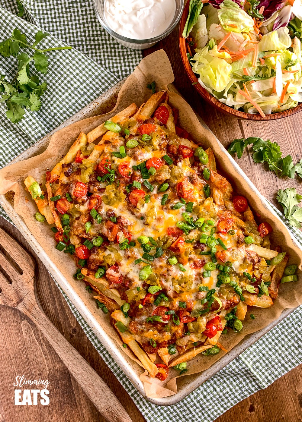 Layered Chilli Cheese Fries on baking tray with salad and soured cream on wooden board with scatter cilatntro