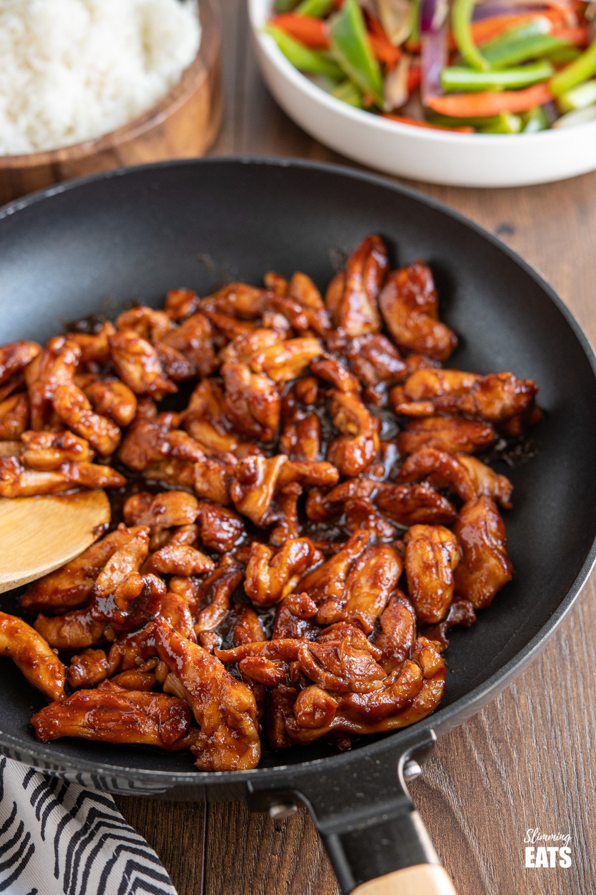 sweet and sticky chicken in wooden handled black frying pan with rice and vegetables in background