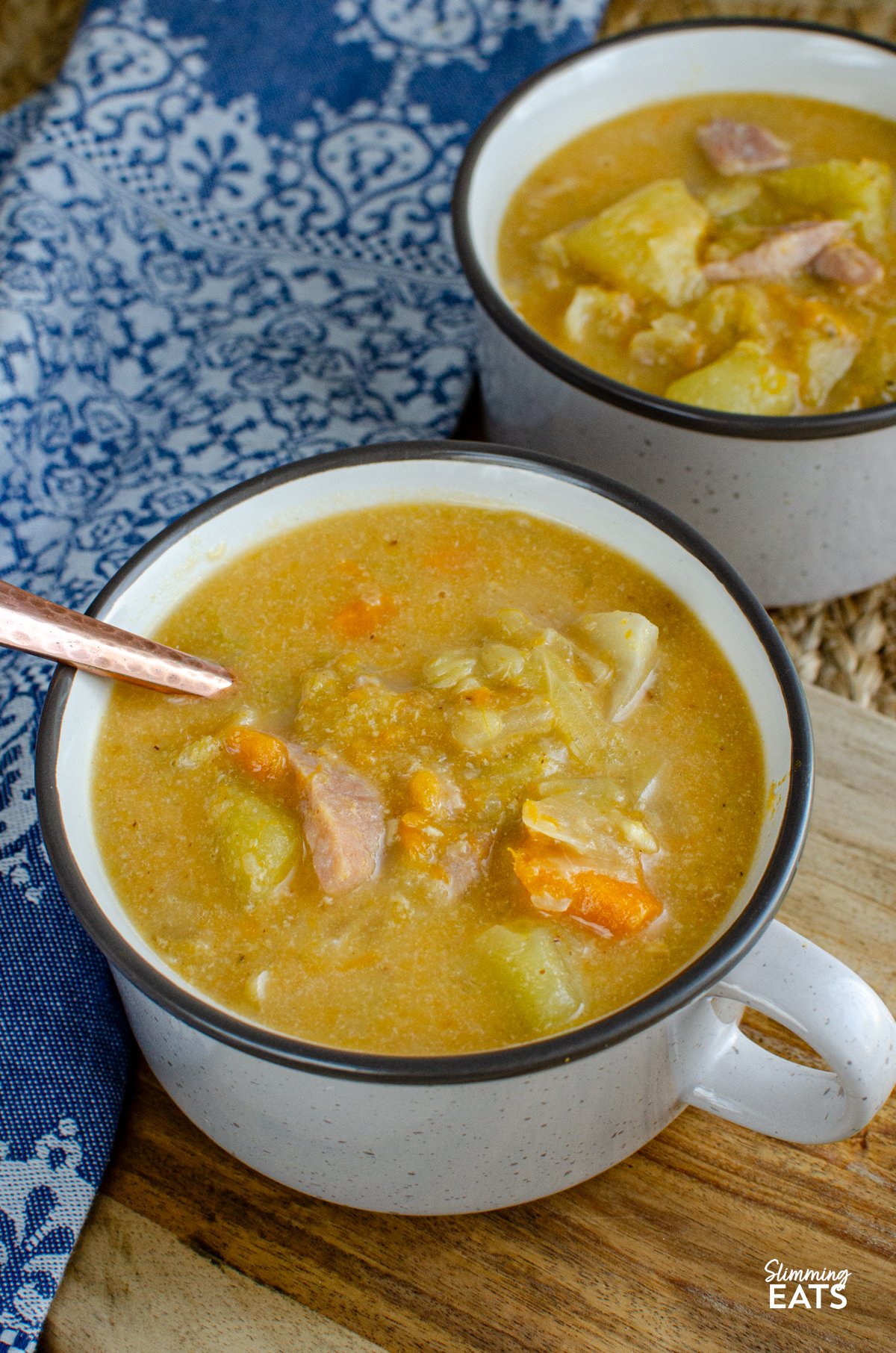 Bacon, Cabbage, and Potato Soup – This hearty soup is a comforting blend of simple ingredients, yet brimming with flavour. The smoky bacon, tender cabbage, and creamy potatoes come together to create a bowl of warmth that’s perfect for any day. Bacon, Cabbage, and Potato Soup servings in two light grey brown soup cup with spoon next to a blue patterned placemat.