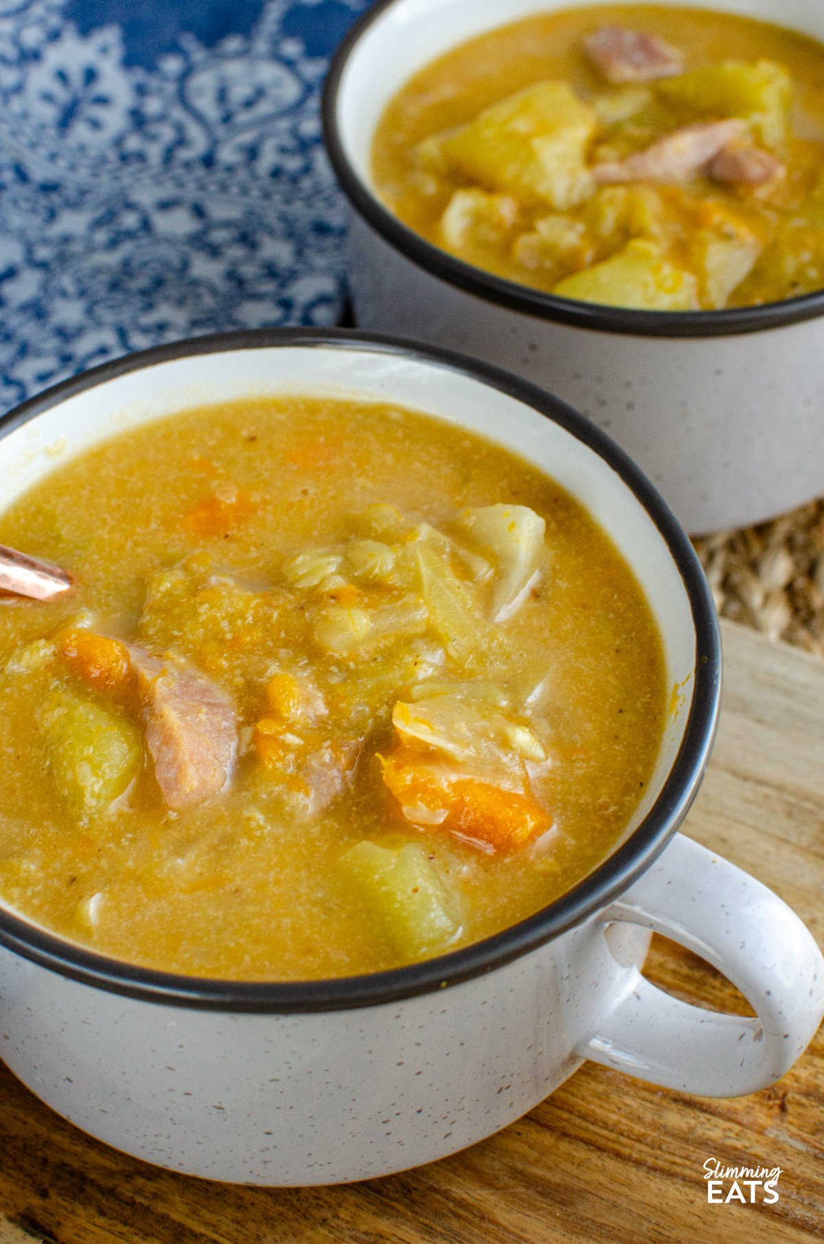 Bacon, Cabbage, and Potato Soup – This hearty soup is a comforting blend of simple ingredients, yet brimming with flavour. The smoky bacon, tender cabbage, and creamy potatoes come together to create a bowl of warmth that’s perfect for any day. Bacon, Cabbage, and Potato Soup servings in two light grey brown soup cup next to a blue patterned placemat.