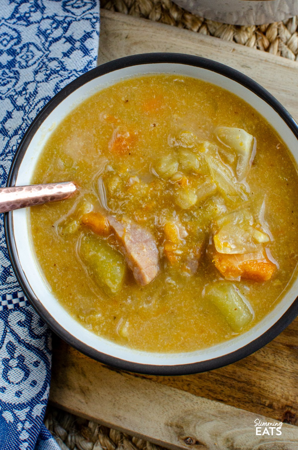 Bacon, Cabbage, and Potato Soup – This hearty soup is a comforting blend of simple ingredients, yet brimming with flavour. The smoky bacon, tender cabbage, and creamy potatoes come together to create a bowl of warmth that’s perfect for any day. close up of Bacon, Cabbage, and Potato Soup servings in light grey brown soup cup next to a blue patterned placemat.