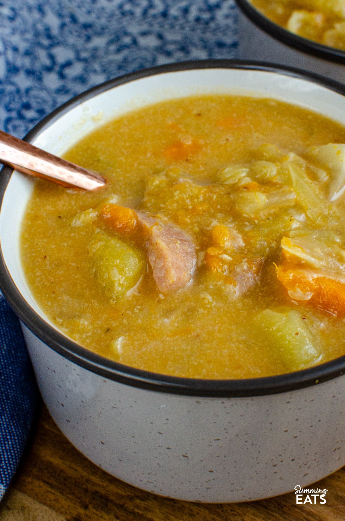Bacon, Cabbage, and Potato Soup – This hearty soup is a comforting blend of simple ingredients, yet brimming with flavour. The smoky bacon, tender cabbage, and creamy potatoes come together to create a bowl of warmth that’s perfect for any day. close up of Bacon, Cabbage, and Potato Soup servings in light grey brown soup cup with spoon next to a blue patterned placemat.