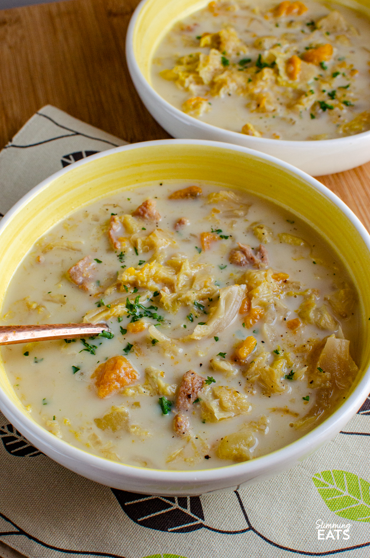 Bacon, Cabbage, and Potato Soup servings in two  white and yellow bowls on a patterned placemat and wooden board