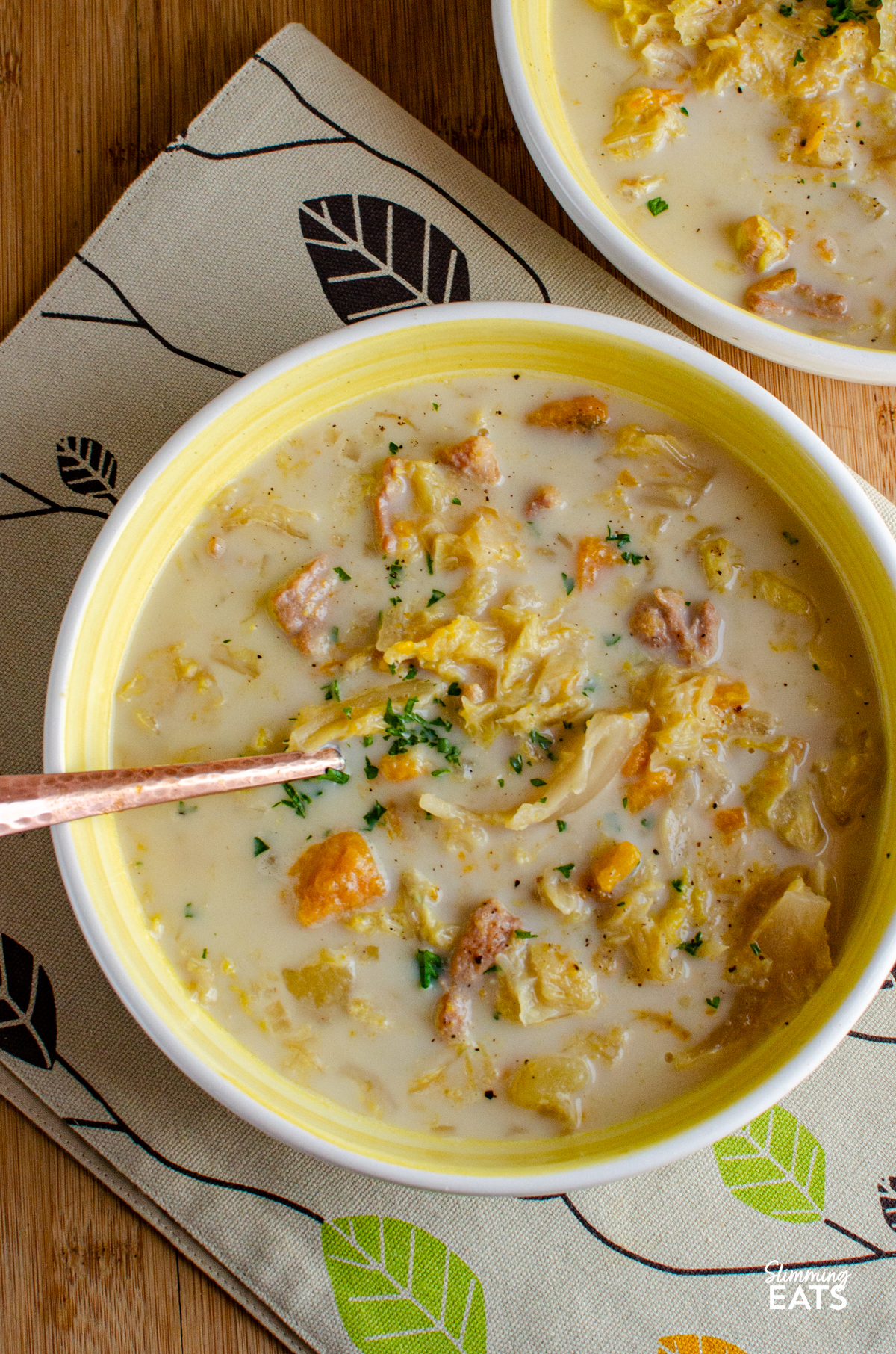 Bacon, Cabbage, and Potato Soup servings in two  white and yellow bowls on a patterned placemat and wooden board