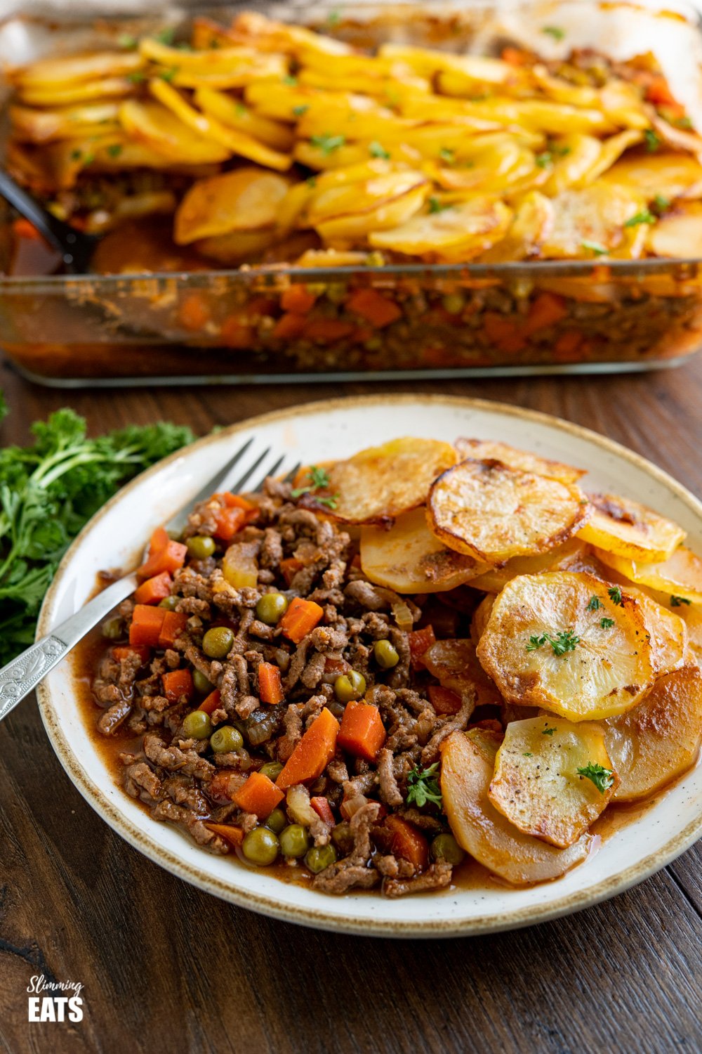 serving of crispy potato topped pie minced beef hotpot on plate with oven proof dish behind