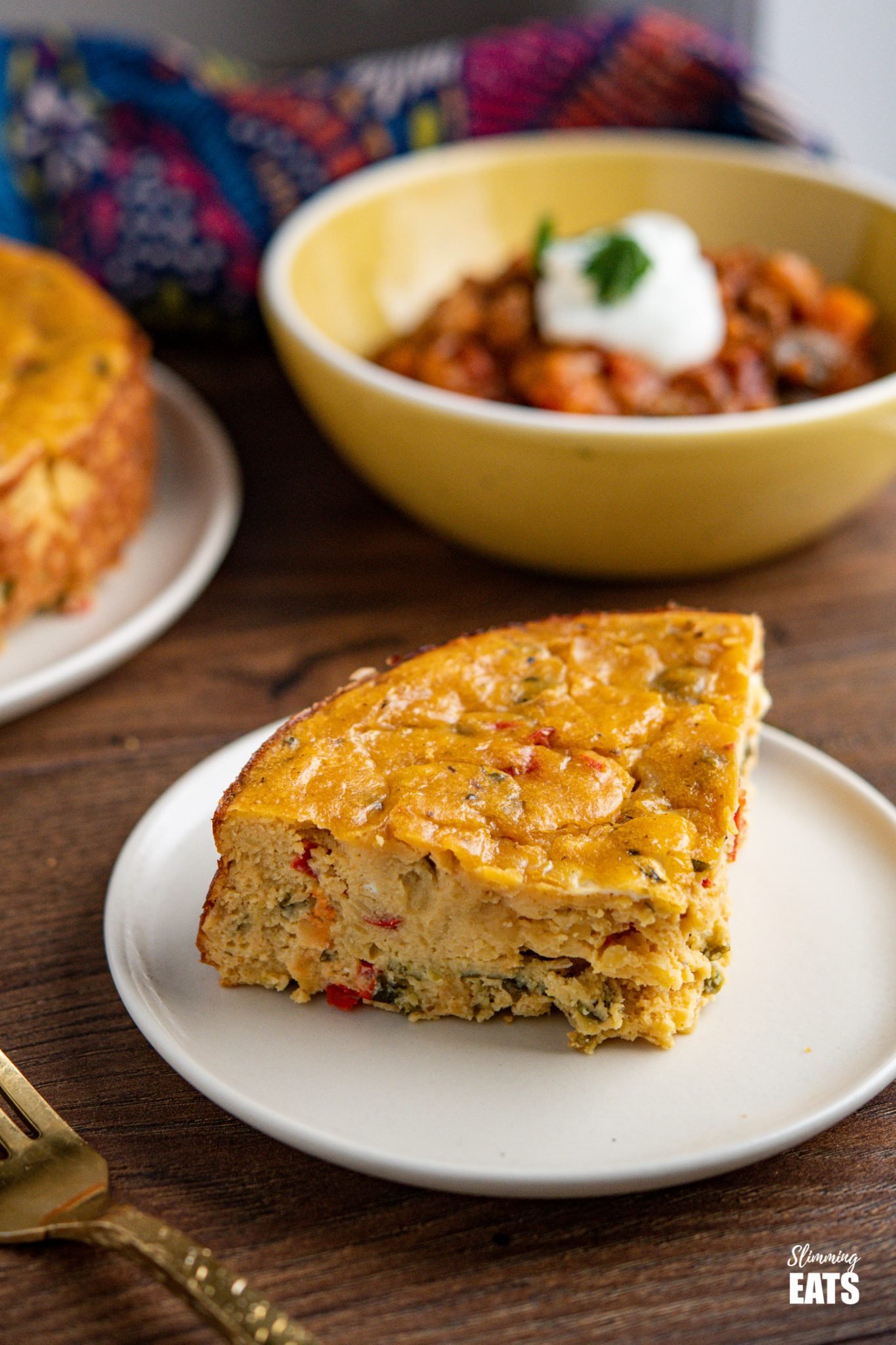 slice of Lentil Cheddar Bake on white plate with yellow bowl of chilli in background 