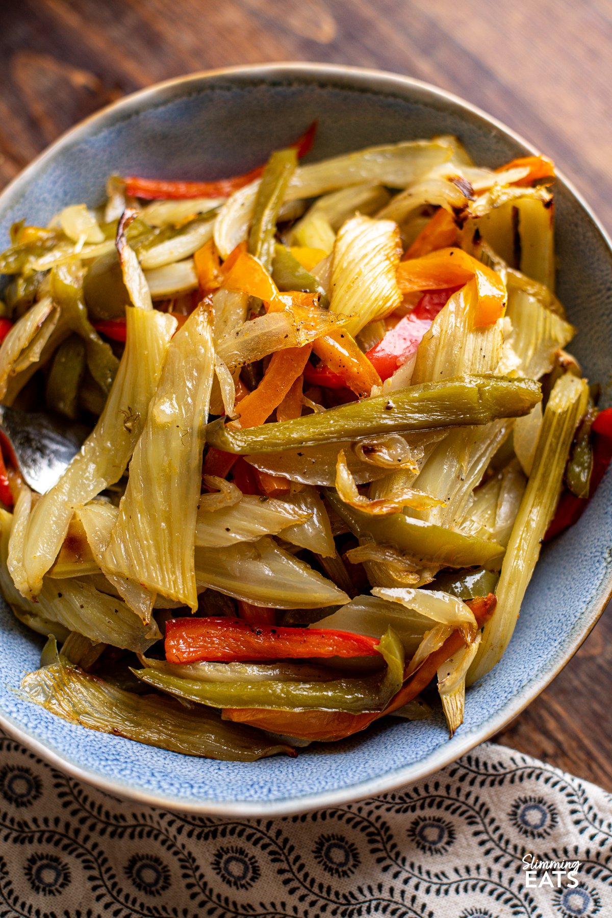 Roasted fennel and peppers is a delicious and nutritious side dish, combining the earthy aniseed flavor of fennel with the sweet essence of bell peppers, all complemented by the aromatic presence of onion and garlic. Roasted Fennel and Mixed Peppers in a blue oval bowl