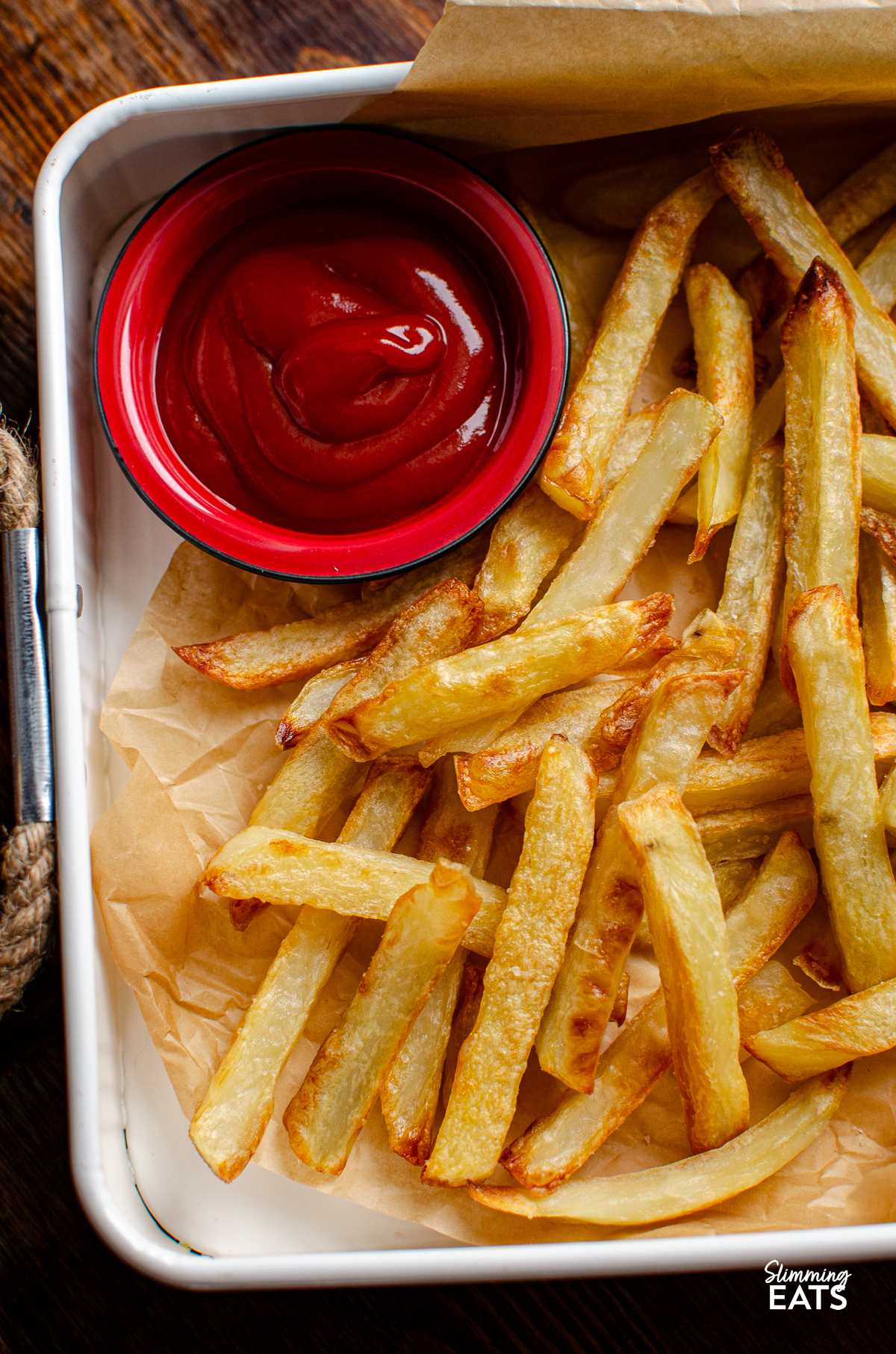 Golden oven baked fries arranged on parchment paper in a white tray with rope handles with a red dish with ketchup