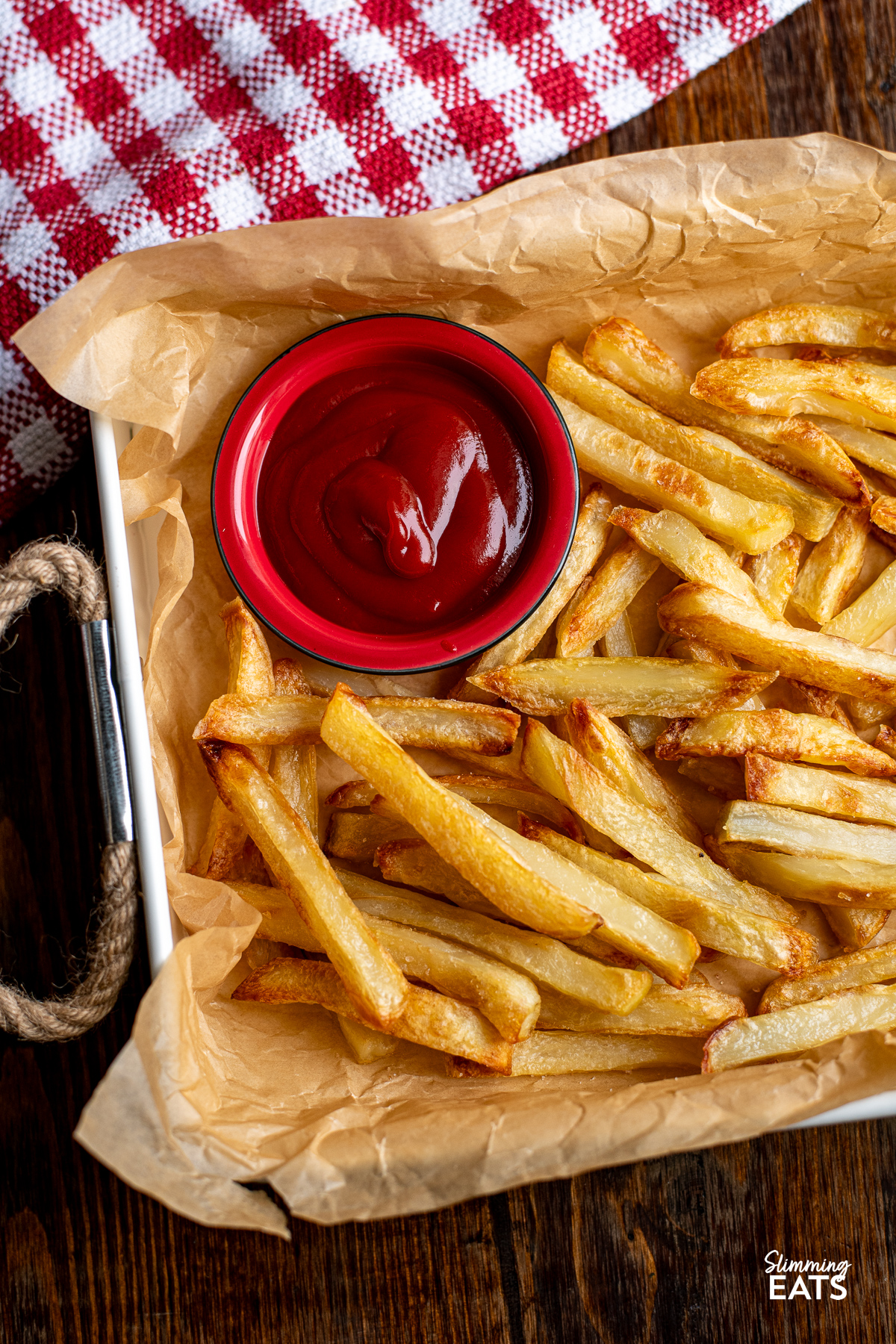 Golden oven baked fries arranged on parchment paper in a white tray with rope handles with a red dish with ketchup