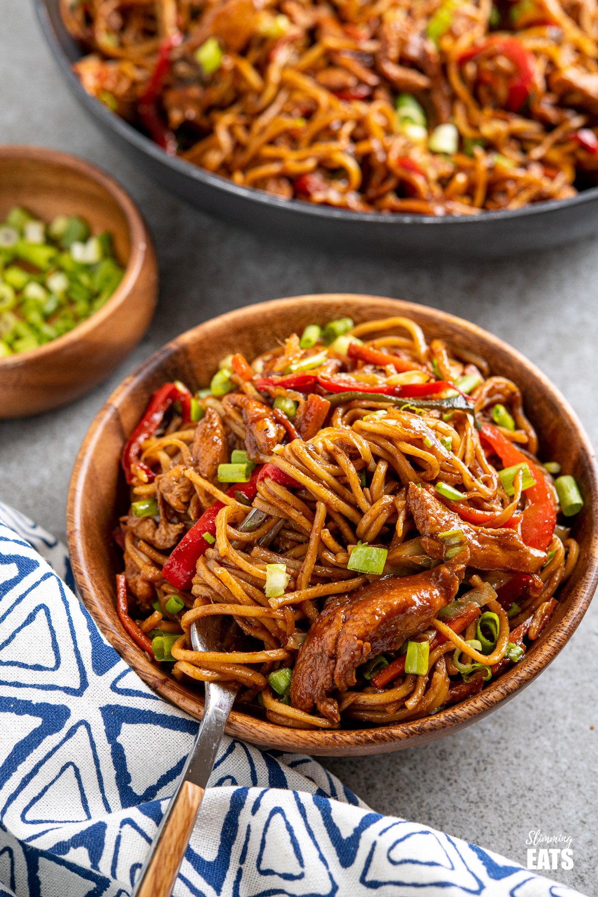 serving of hoisin chicken noodles in wooden bowl with fork place to left in bowl and frying pan of noodles in background