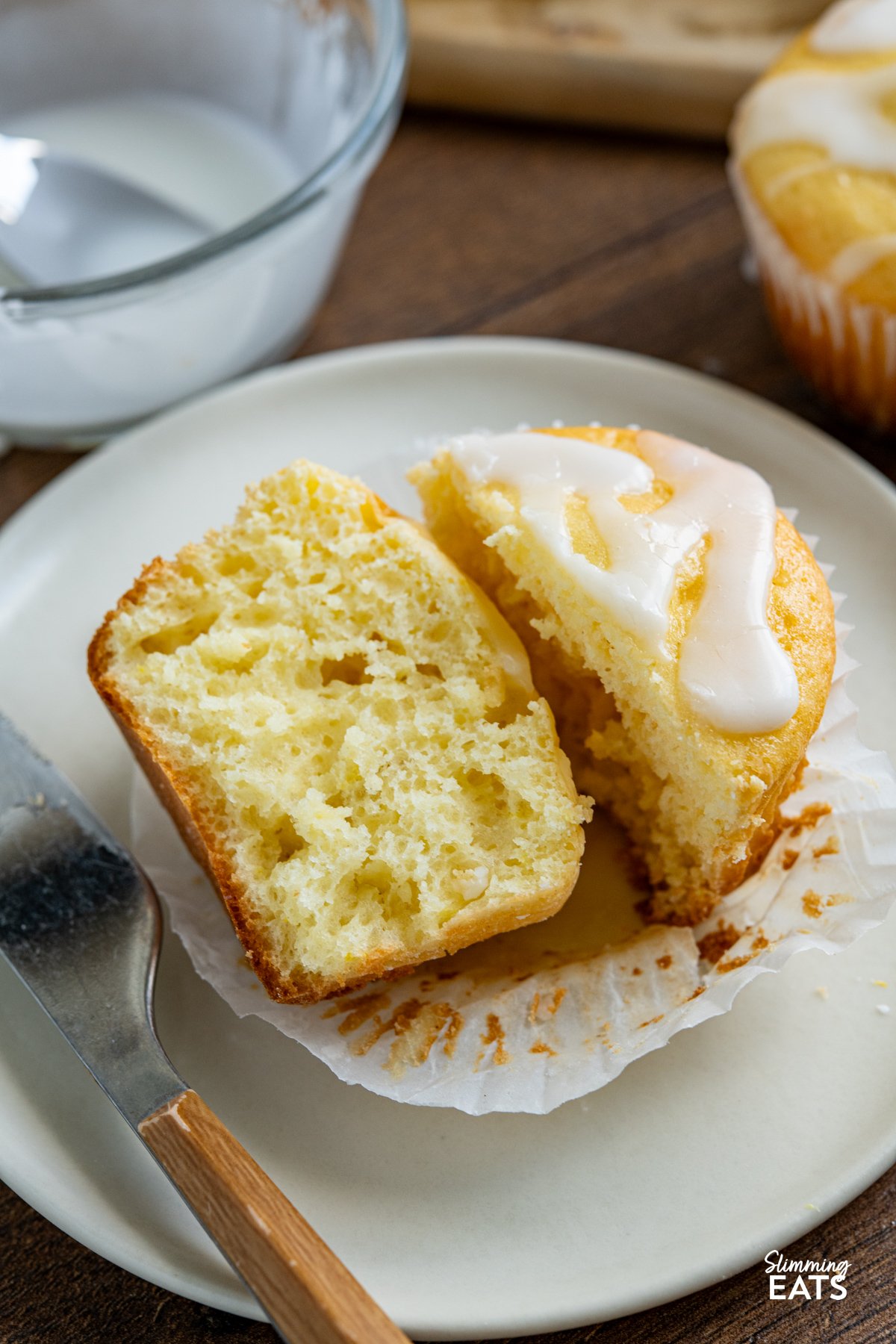 These Lemon Drizzle Muffins are a light and zesty treat that deliver all the flavour of a classic bake without the extra calories. Lemon Drizzle Muffins sliced in half on a plate with a knife next to it.