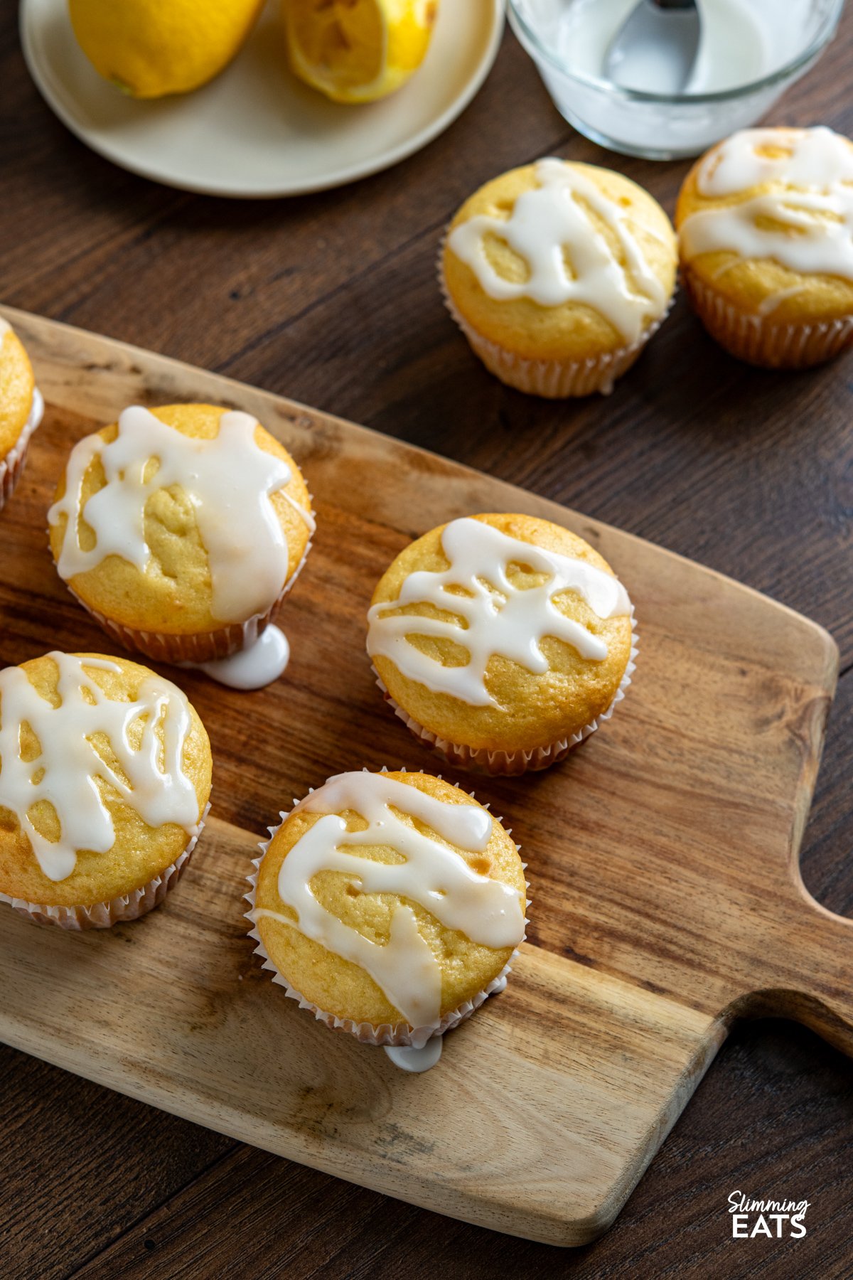 These Lemon Drizzle Muffins are a light and zesty treat that deliver all the flavour of a classic bake without the extra calories. Lemon Drizzle Muffins on a wooden board with a bowl of icing with a spoon