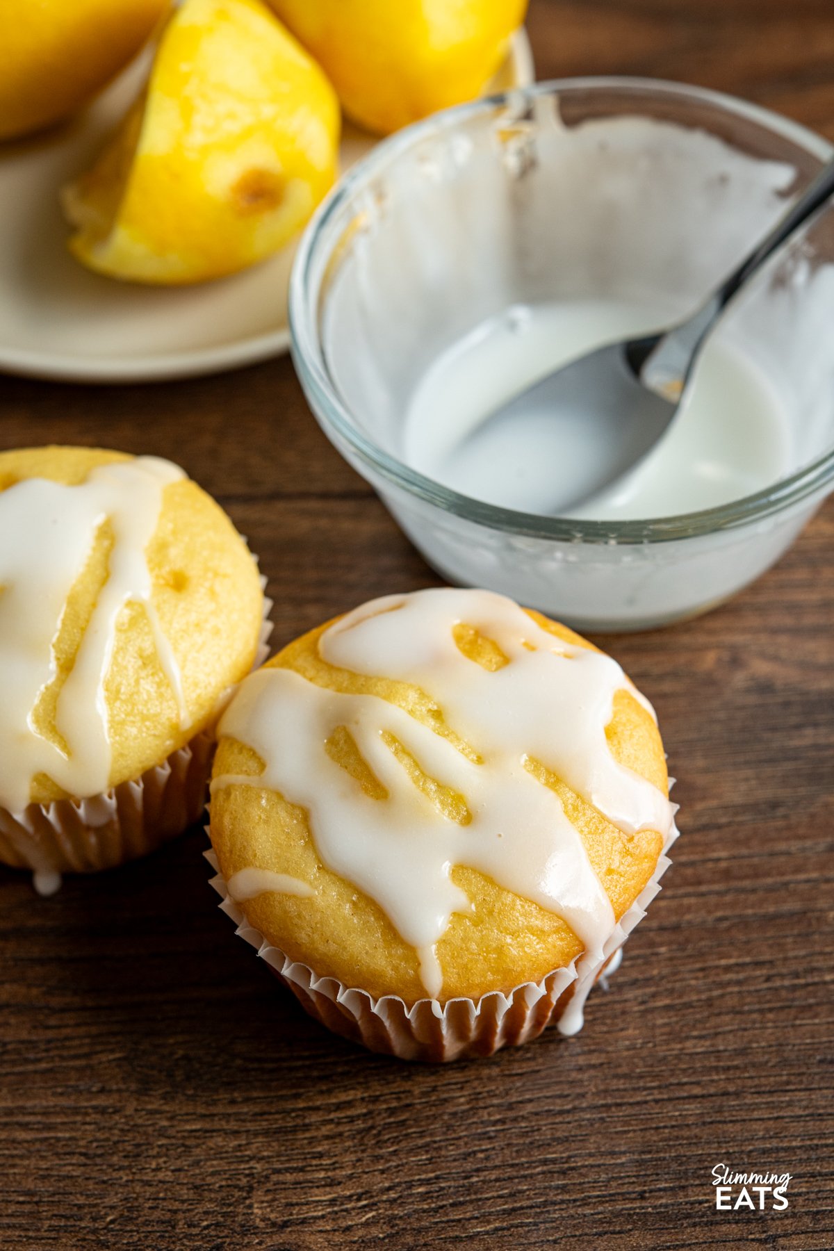 These Lemon Drizzle Muffins are a light and zesty treat that deliver all the flavour of a classic bake without the extra calories. two Lemon Drizzle Muffins with bowl of icing and spoon next to them.