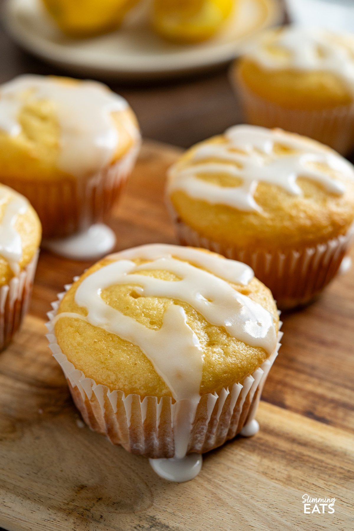 These Lemon Drizzle Muffins are a light and zesty treat that deliver all the flavour of a classic bake without the extra calories. close up of Lemon Drizzle Muffins on a board with plate of cut lemons and bowl of icing in background