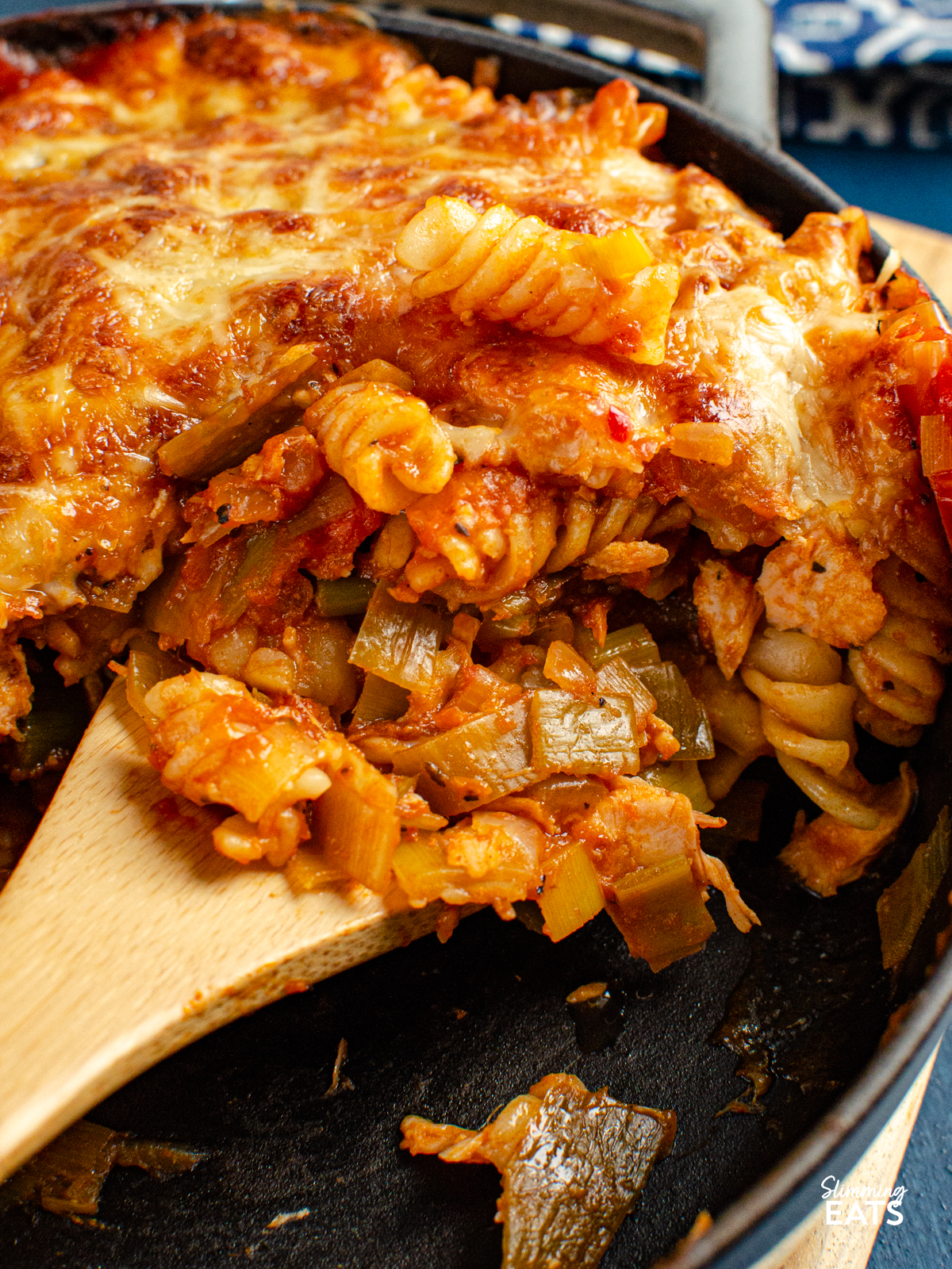 Salmon and Leek Pasta Bake - Salmon and Leek Pasta Bake - a delicious recipe using fresh salmon, sauteed leeks and a homemade tomato pasta sauce, all finished off with melted cheesy goodness - yum!! Slimming Eats and Weight Watchers friendly close up of wooden spoon scooping up Salmon and Leek Pasta Bake from grey cast iron Staub skillet